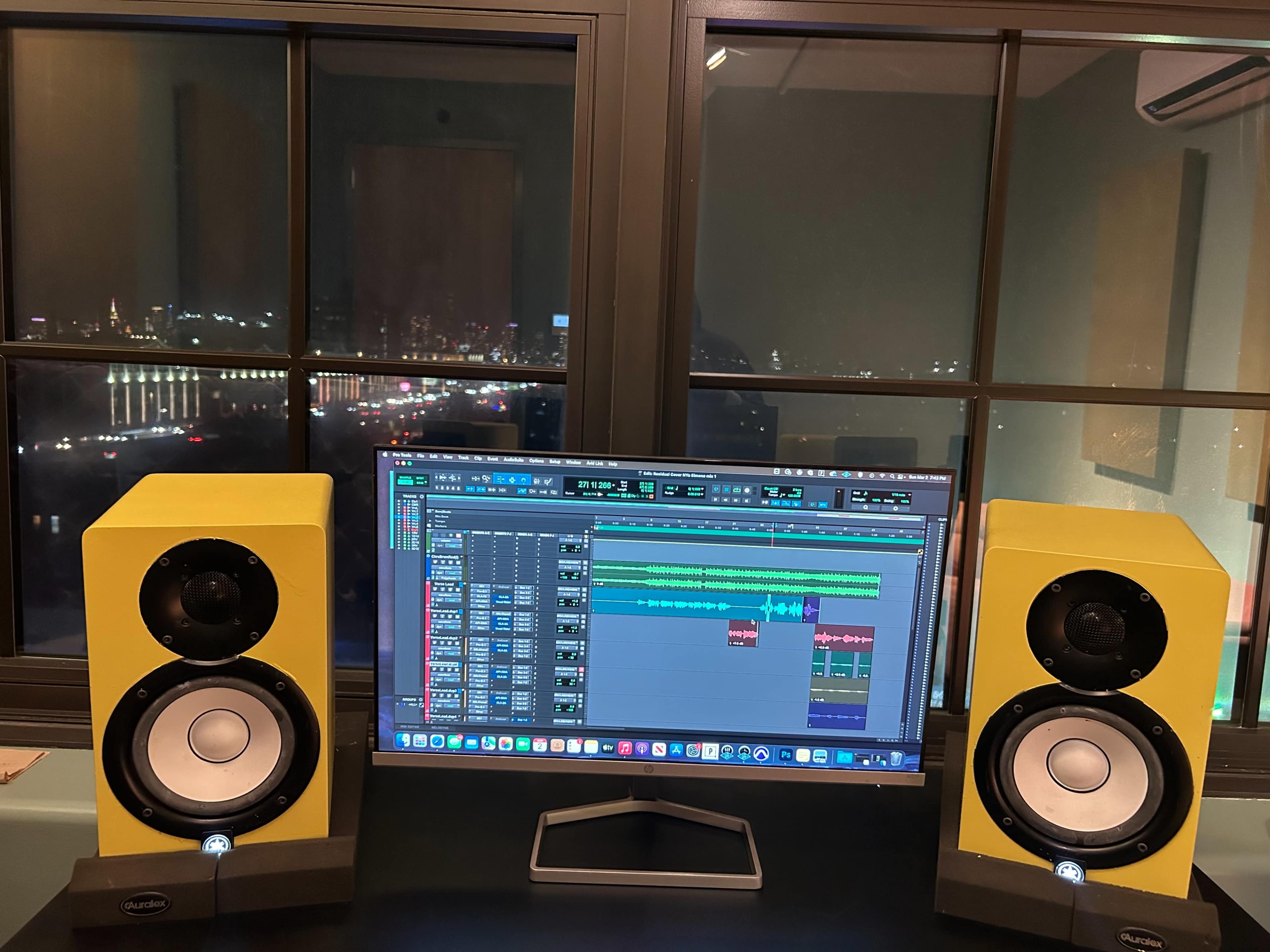 A computer monitor displaying a music editing software is positioned between two yellow studio monitors, with a city skyline visible through the window in the background.