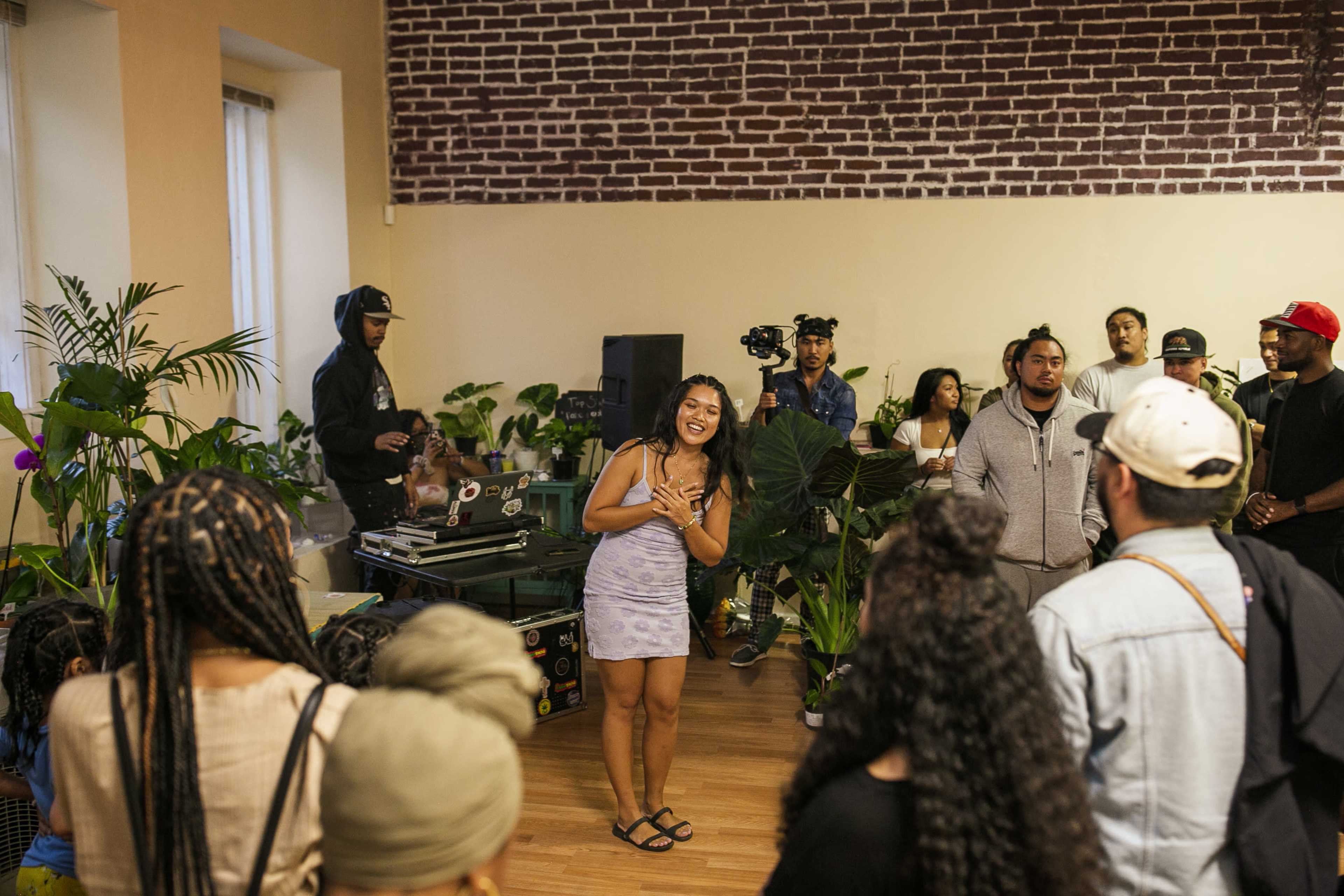 A woman stands in the center of a room filled with plants, engaging a crowd while a DJ plays music in the background.