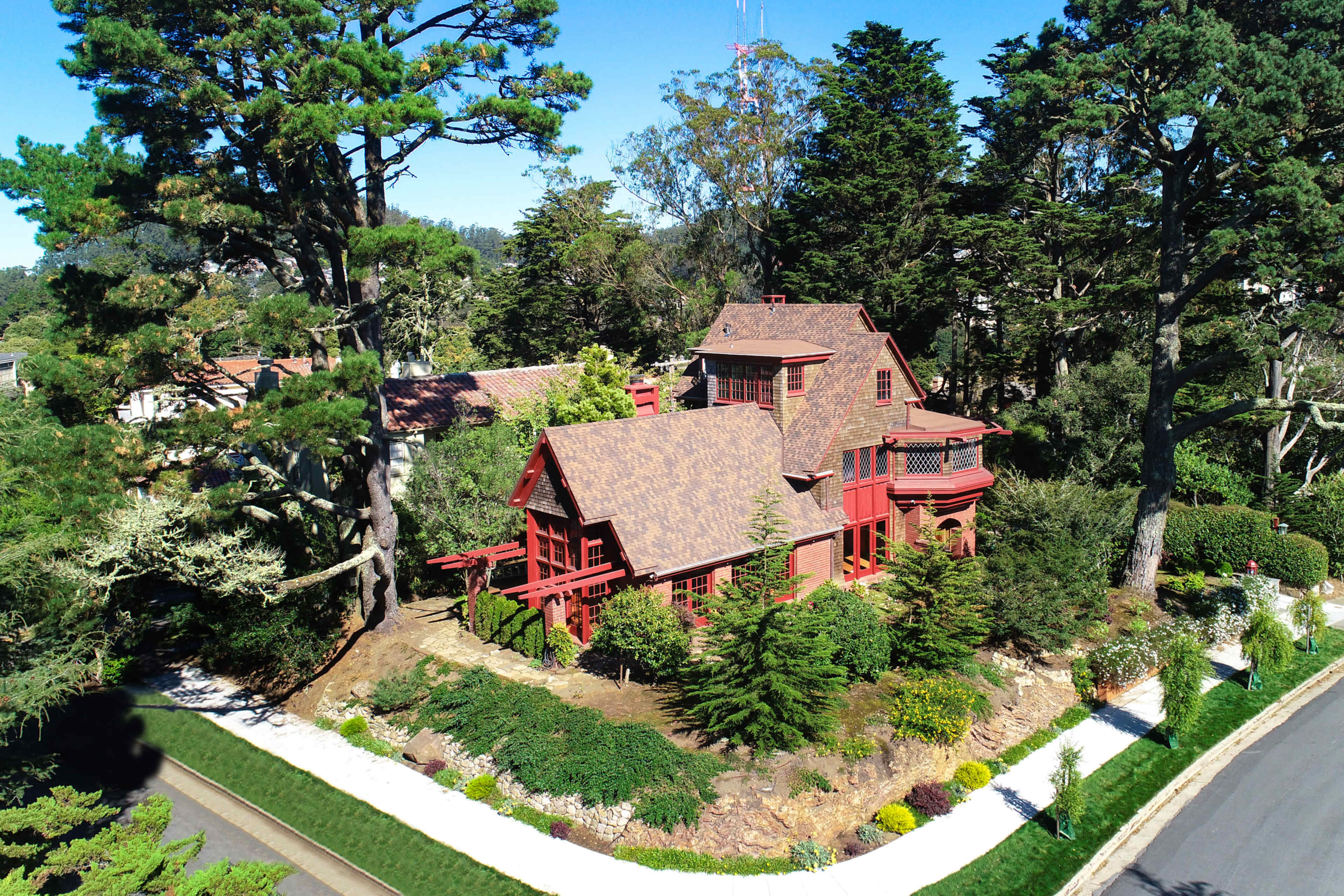 A red, wooden house is surrounded by trees and landscaping on a corner lot near a road.