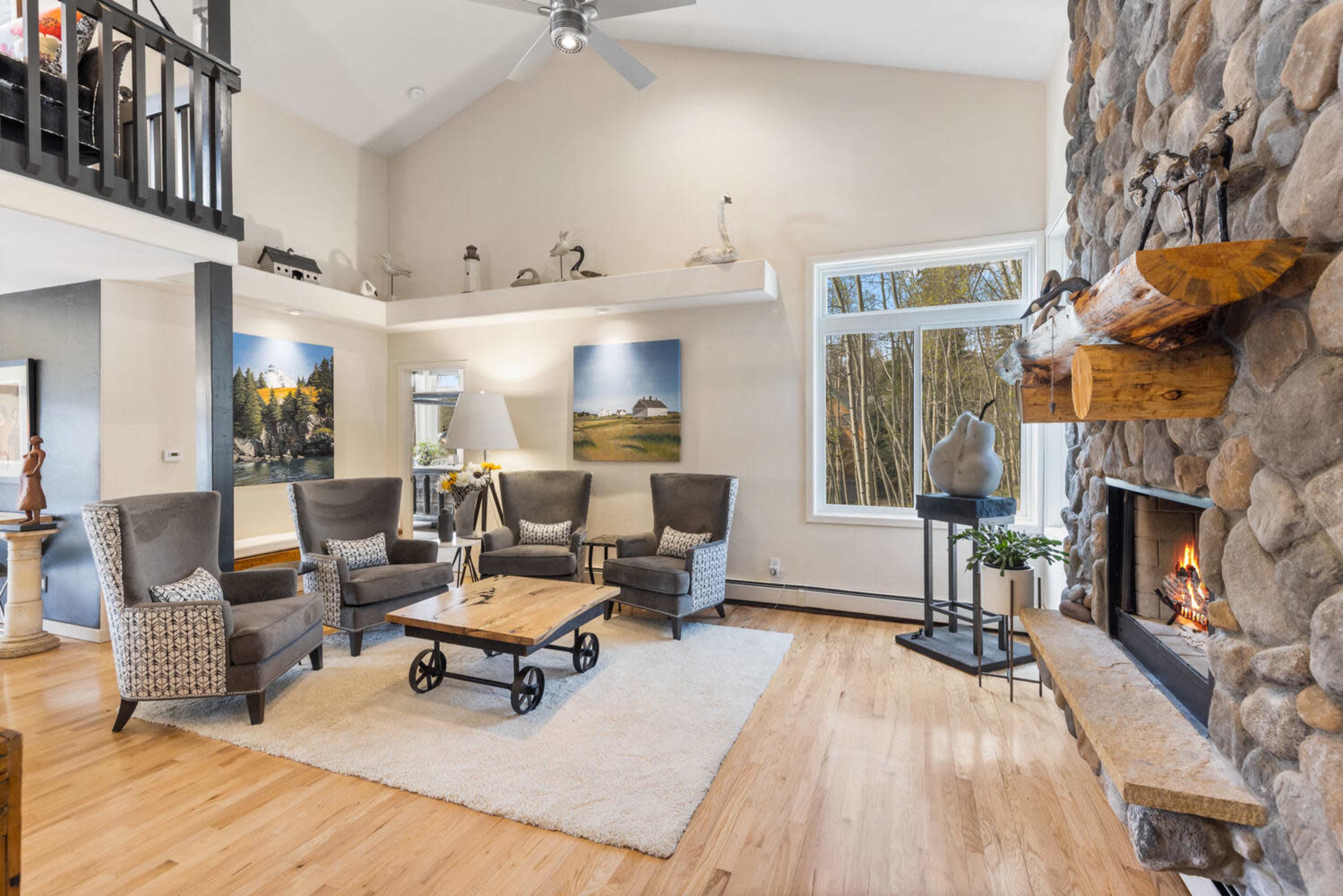 The image shows a well-lit living room featuring a stone fireplace, comfortable seating arrangements, and large windows facing a wooded view.