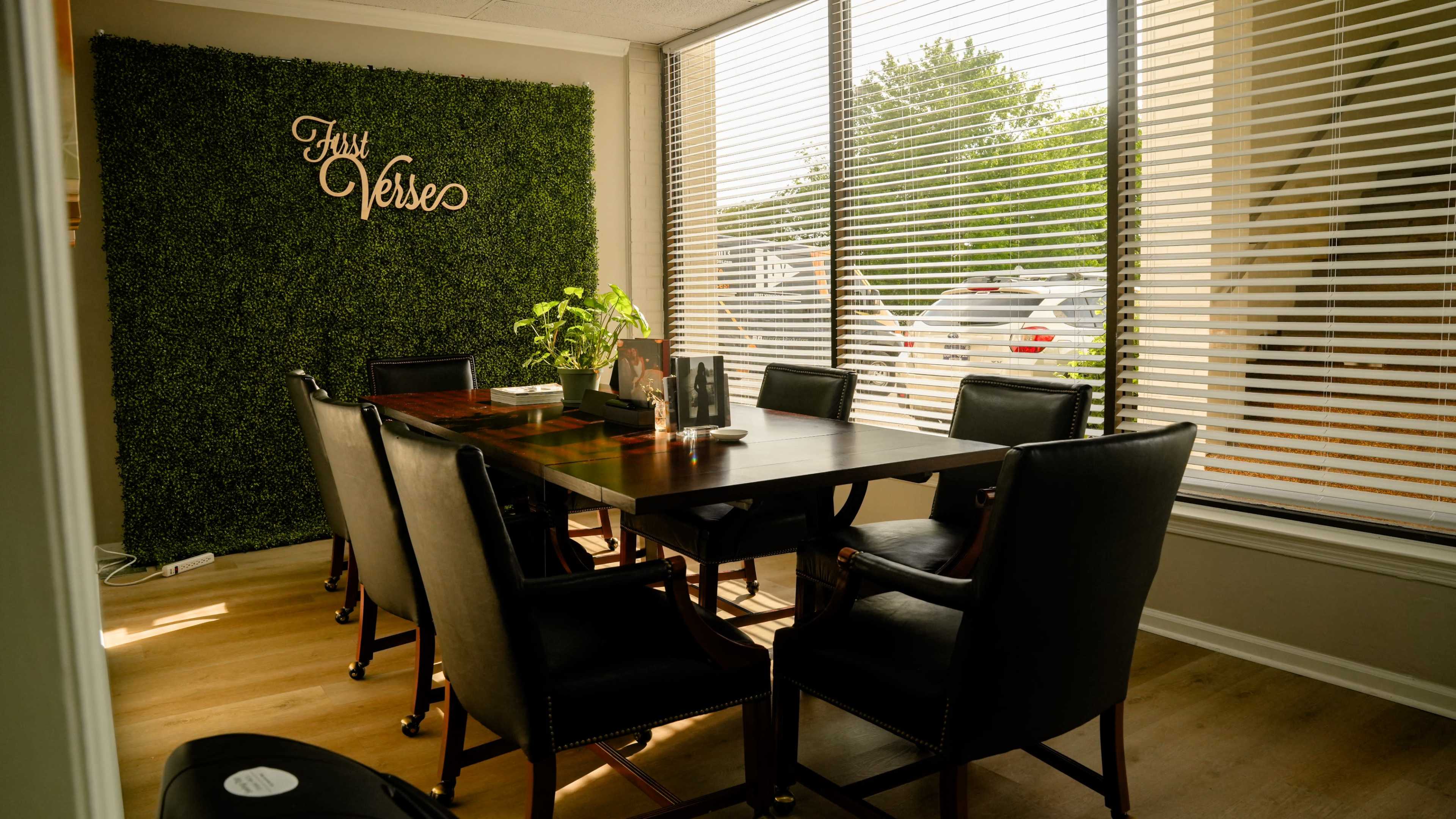 A conference room features a large wooden table surrounded by black leather chairs, with a wall covered in greenery and a sign reading "Just Verse."