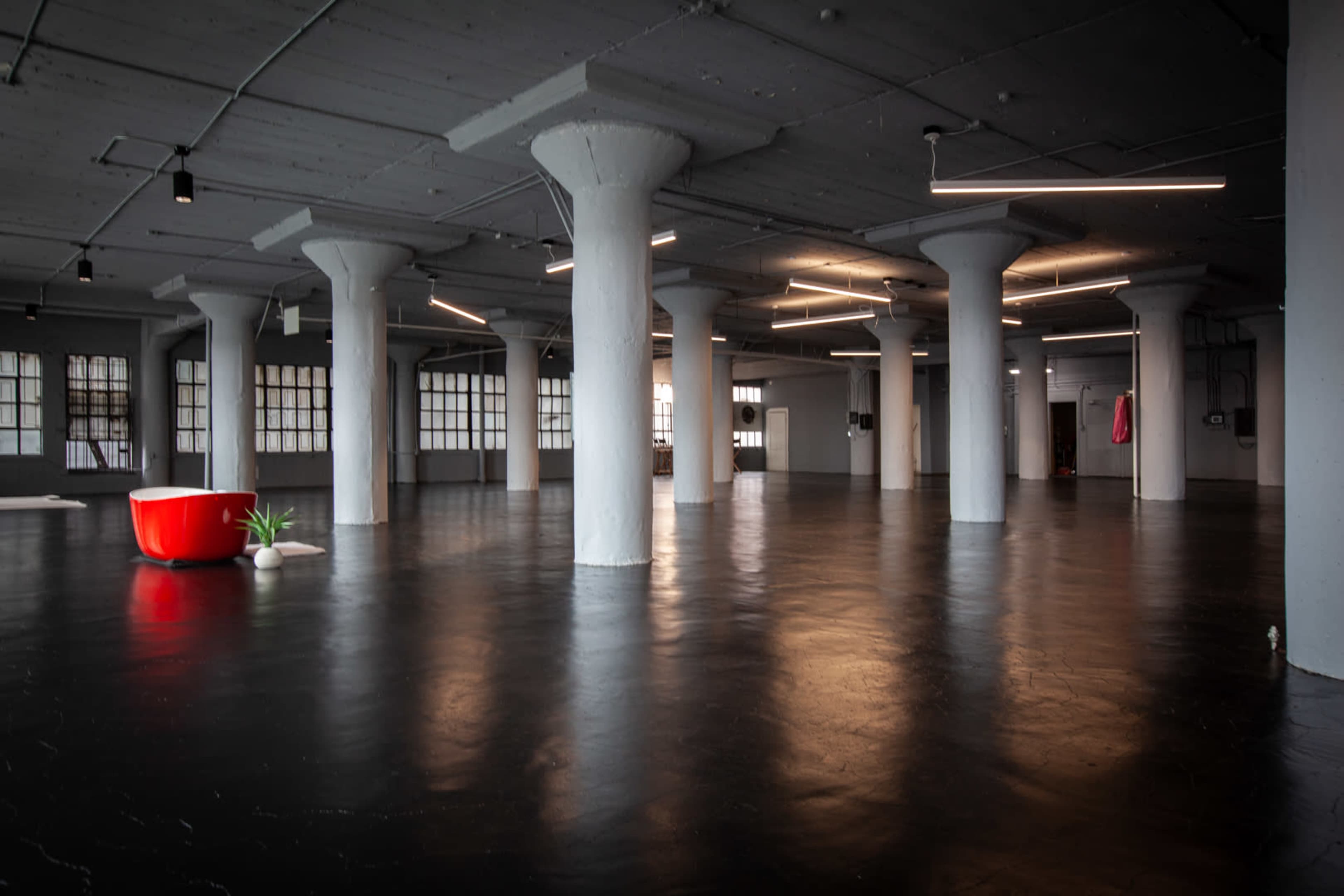 The image shows a spacious industrial-style interior with polished black floors, tall white columns, and several overhead light fixtures, featuring a red bathtub and a small plant in one corner.