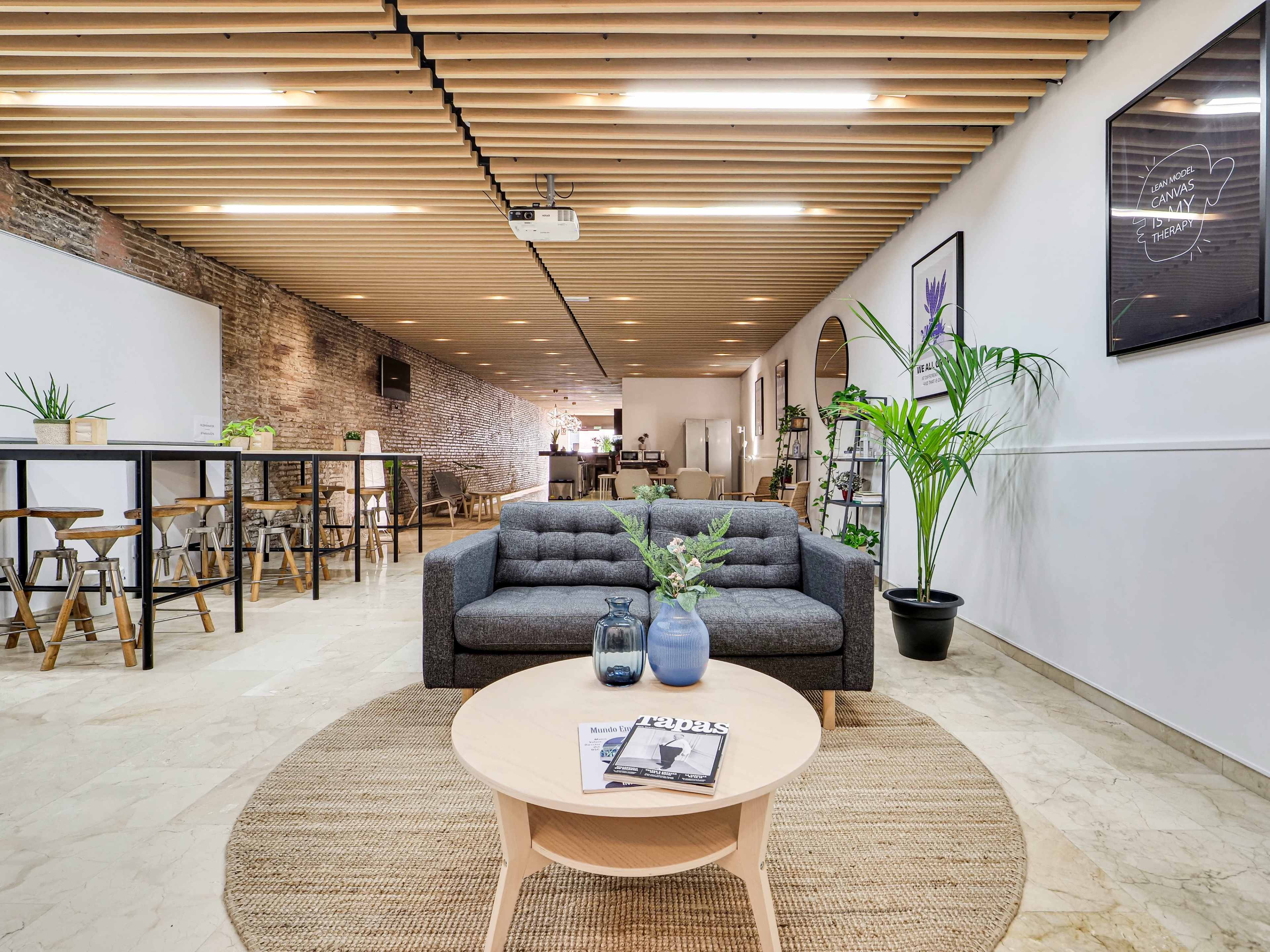 The image shows a modern interior space featuring a gray sofa, a round coffee table with a magazine on it, and a plant, set against a backdrop of exposed brick walls and wooden ceiling beams.