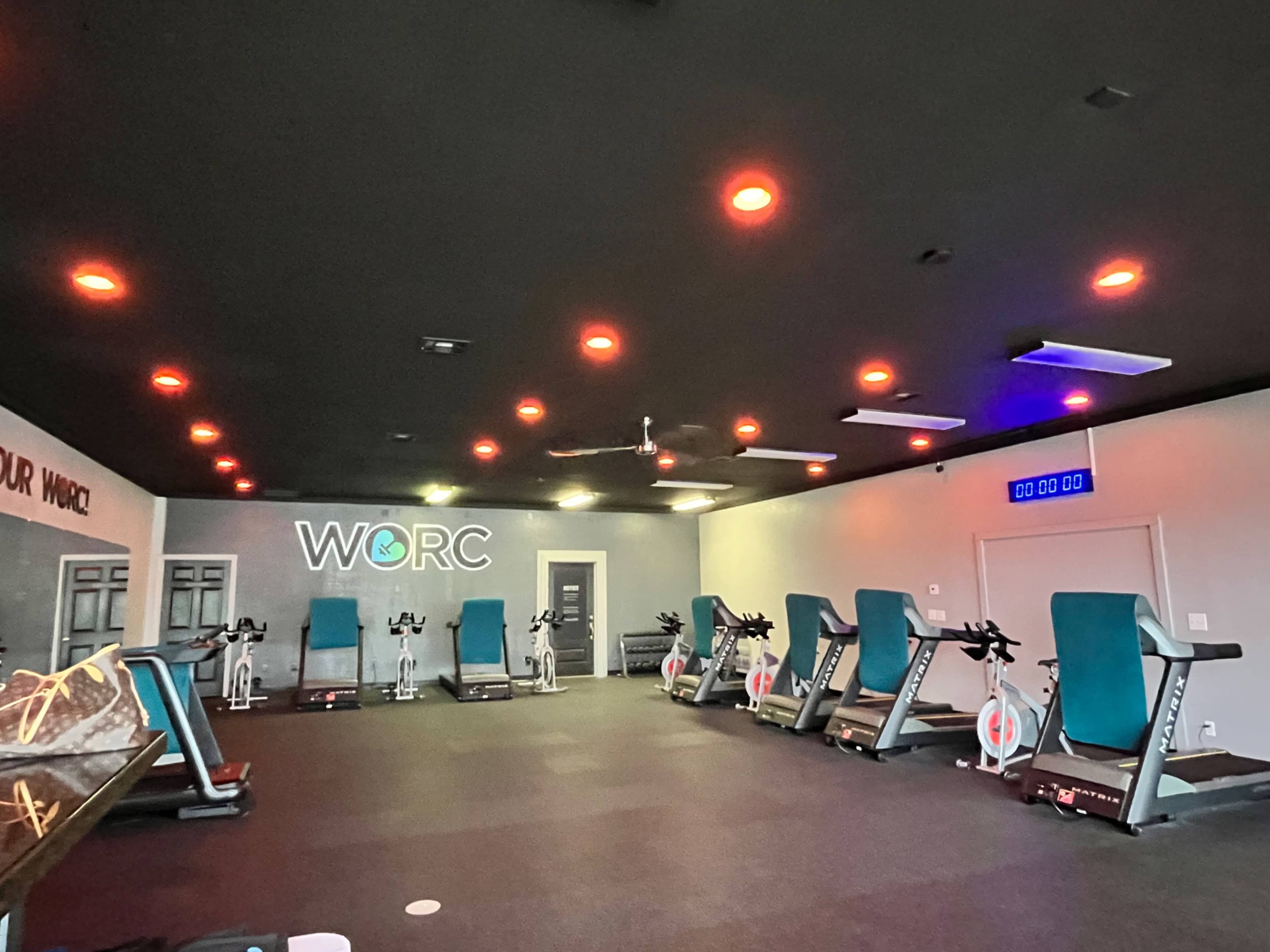 The image shows a gym interior featuring several exercise bikes arranged in a spacious room with dim lighting and a large "WORC" logo on the wall.