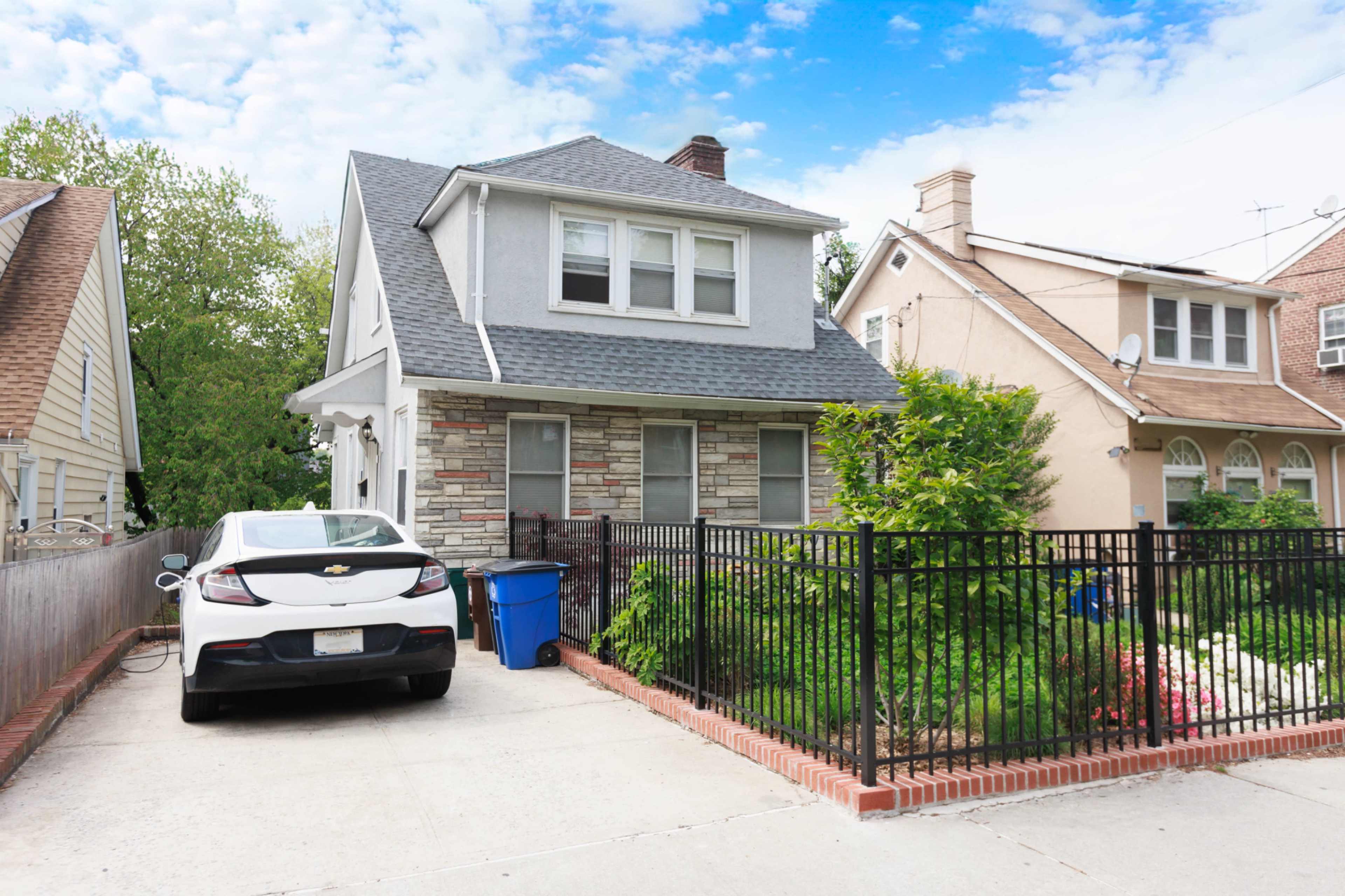 A two-story house with a stone façade and a gray roof sits next to a parked car in a driveway, surrounded by greenery and a black metal fence.