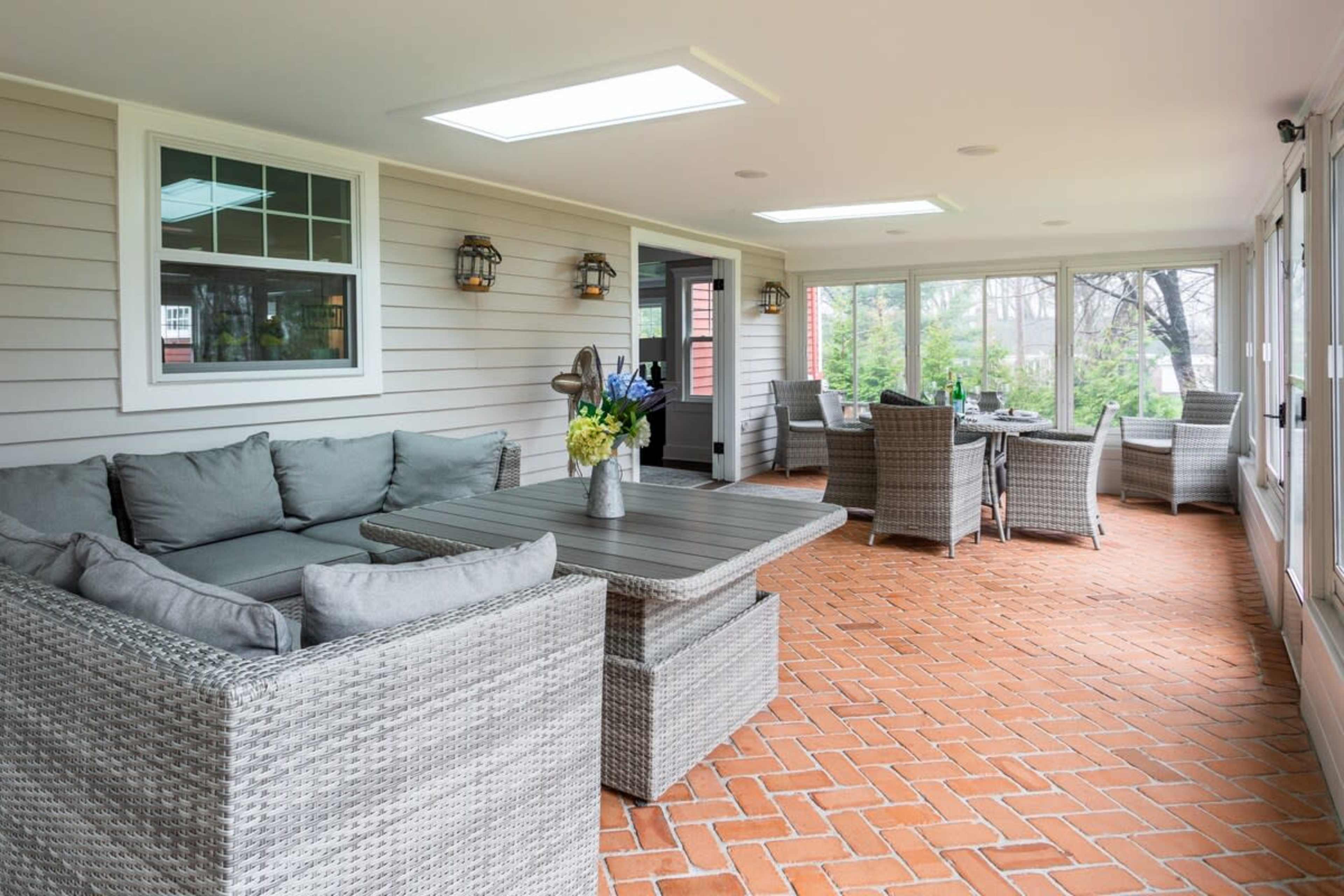 A sunlit enclosed porch with comfortable seating and a table, featuring brick flooring and large windows.