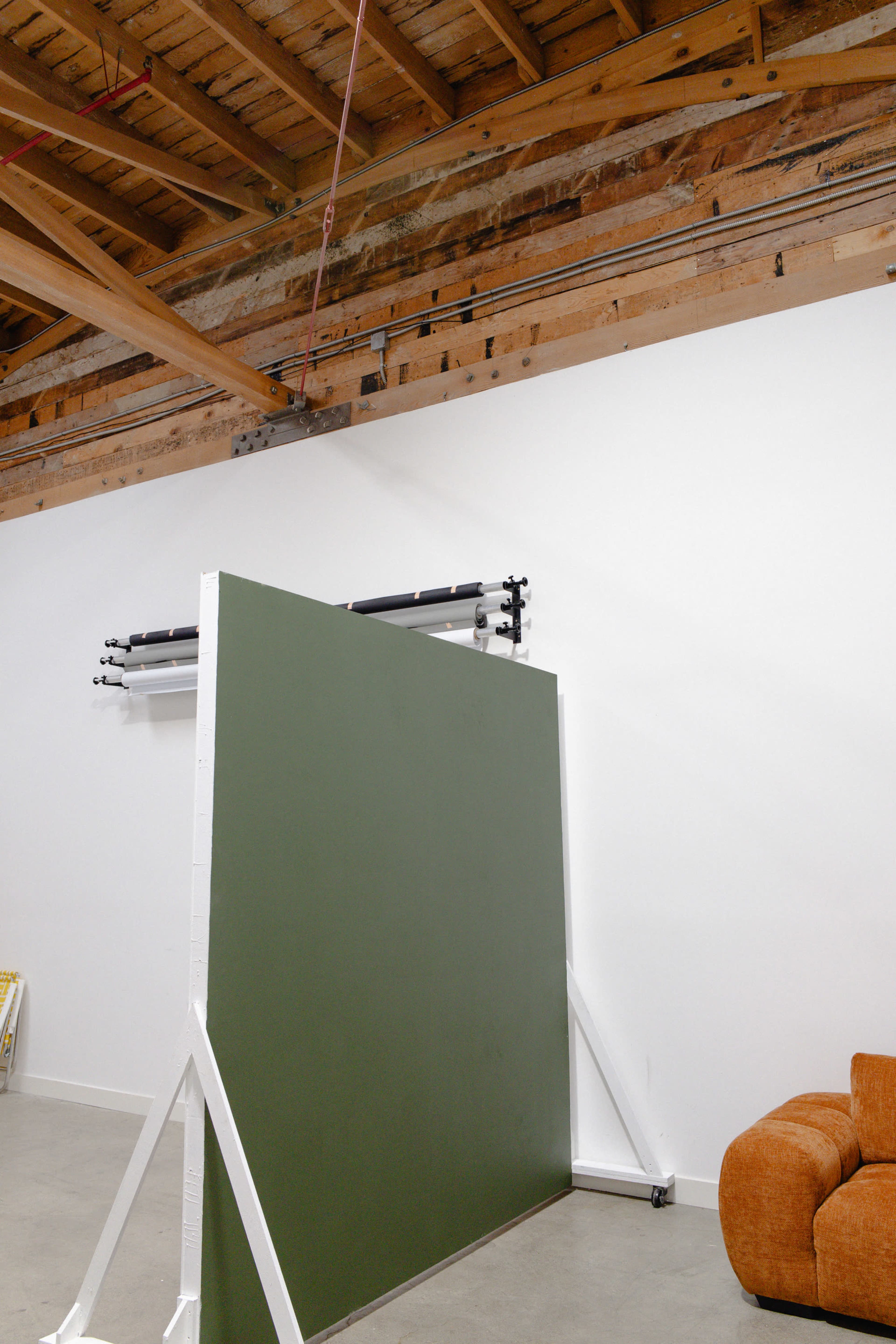 A green backdrop on a stand is positioned in a spacious room with wooden beams and a beige sofa.