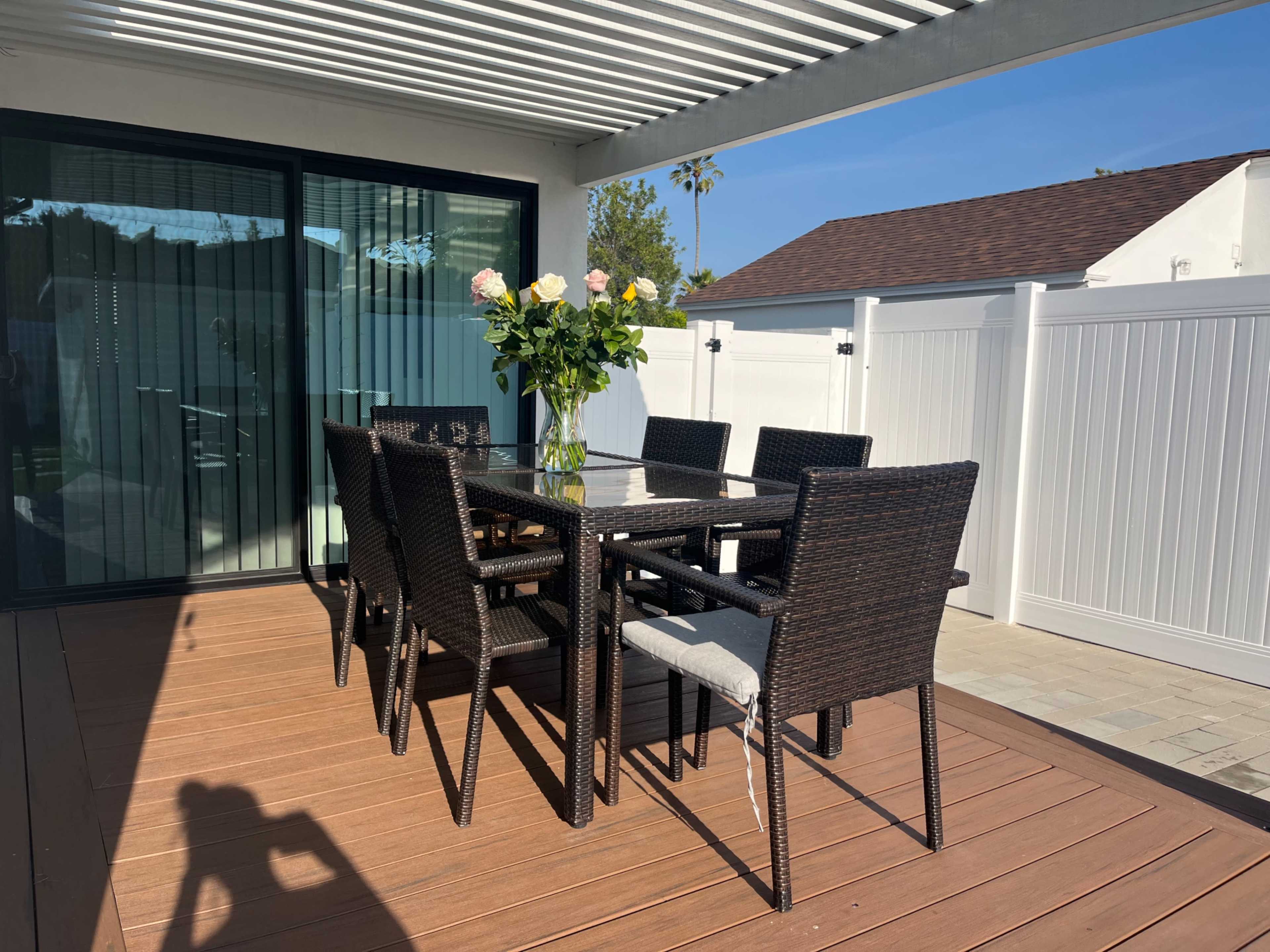 A glass-top dining table is set with six black wicker chairs under a pergola on a wooden deck, with a vase of roses in the center.