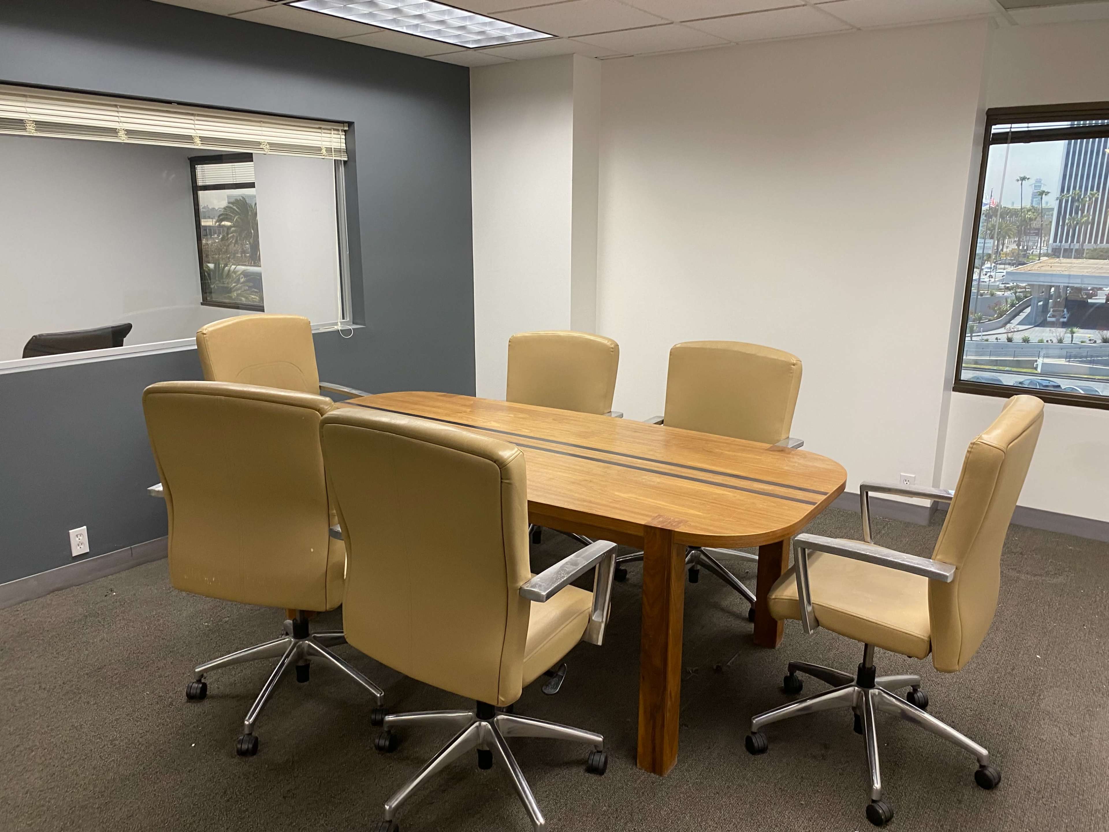 A conference room features a wooden table surrounded by six leather office chairs, with a large window showing an exterior view.