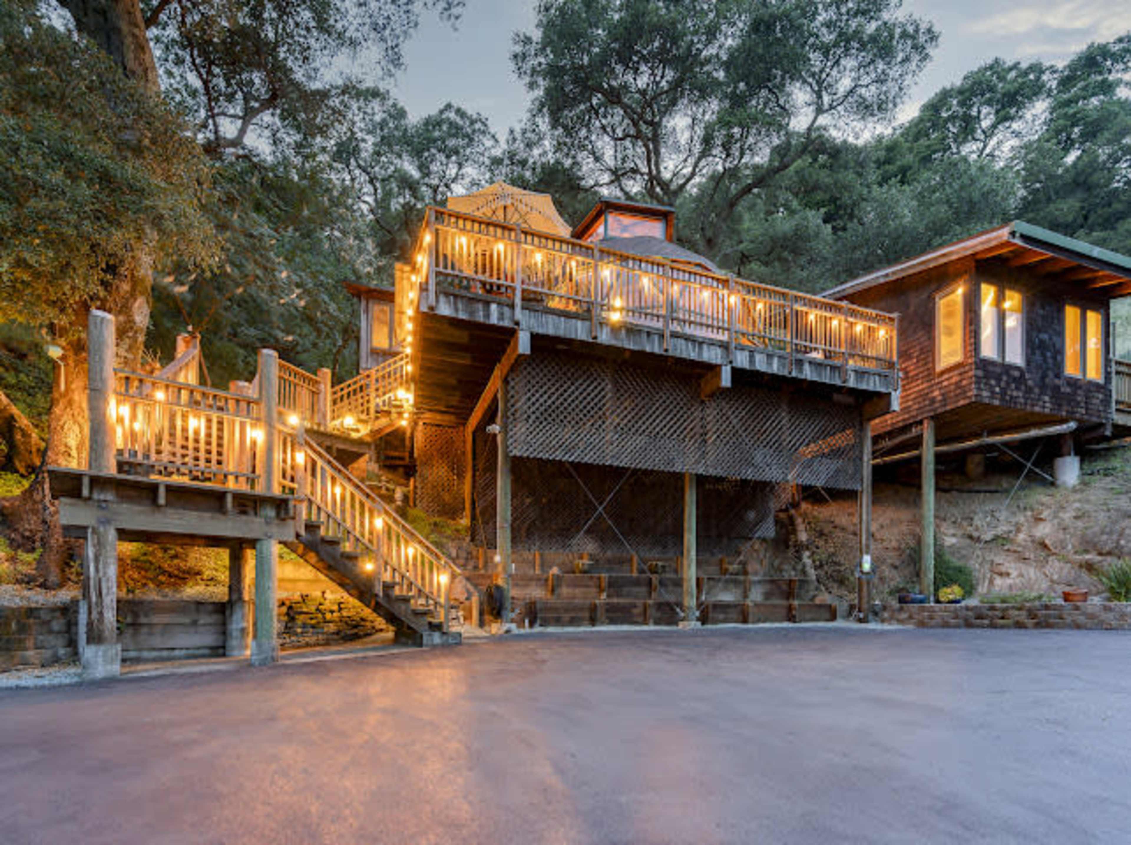 A wooden deck with string lights surrounds a multi-level house set on a hillside, with a staircase leading up to the entrance.