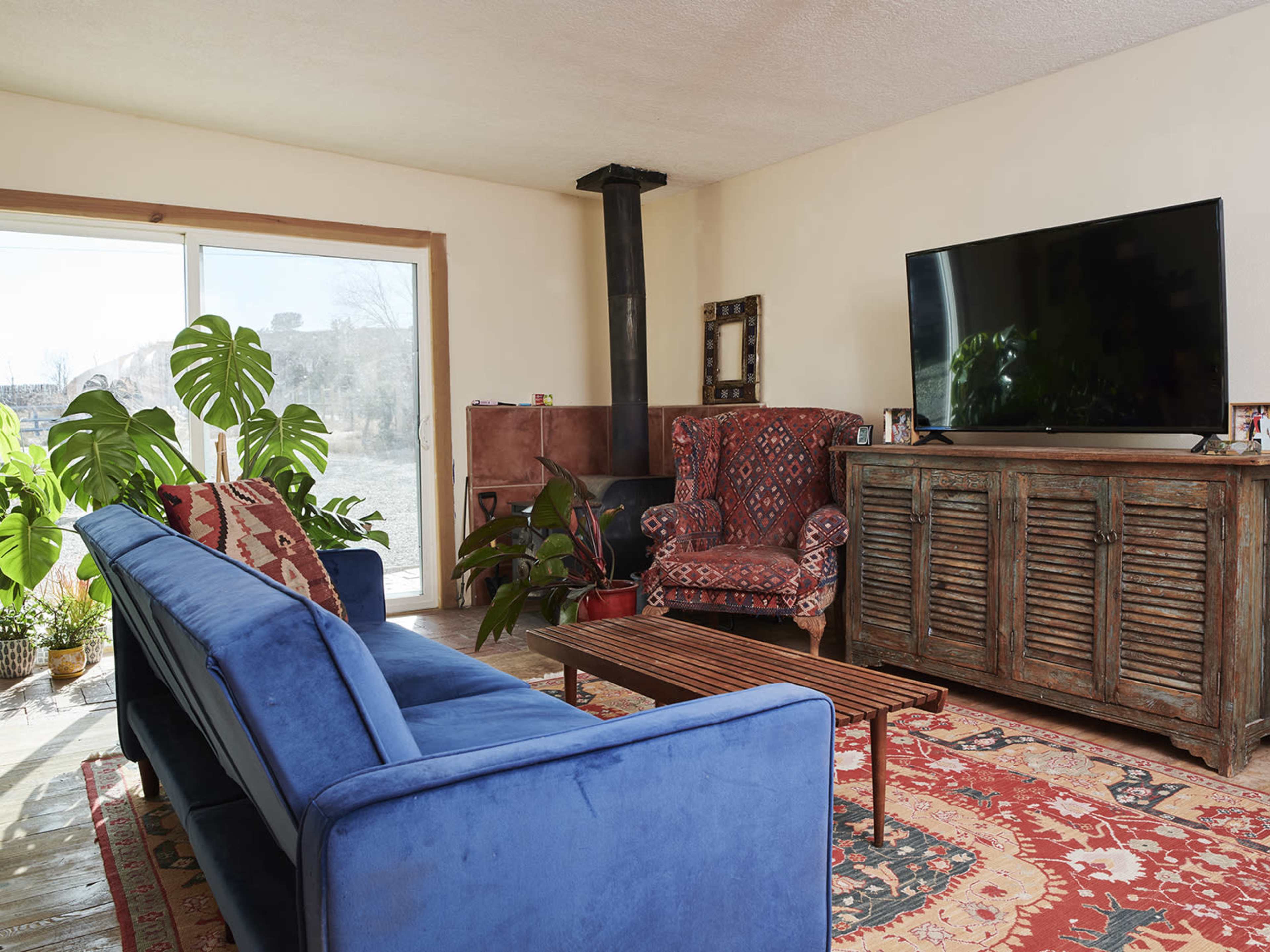 The living room features a blue couch, a patterned armchair, a wooden coffee table, and a television on a rustic cabinet, with large windows letting in natural light.