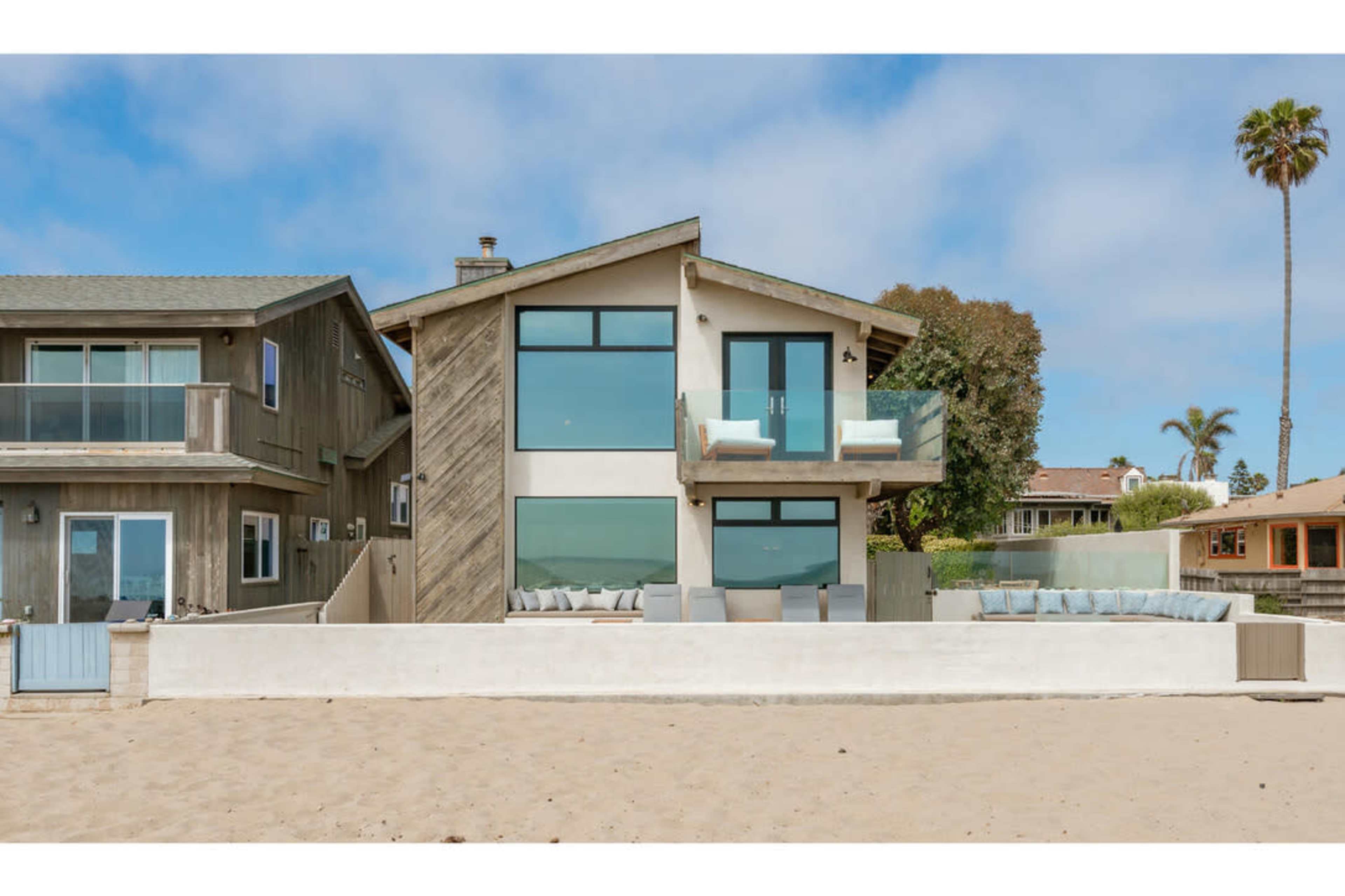 A modern beach house with large windows and a balcony stands along a sandy shore, adjacent to two neighboring homes.