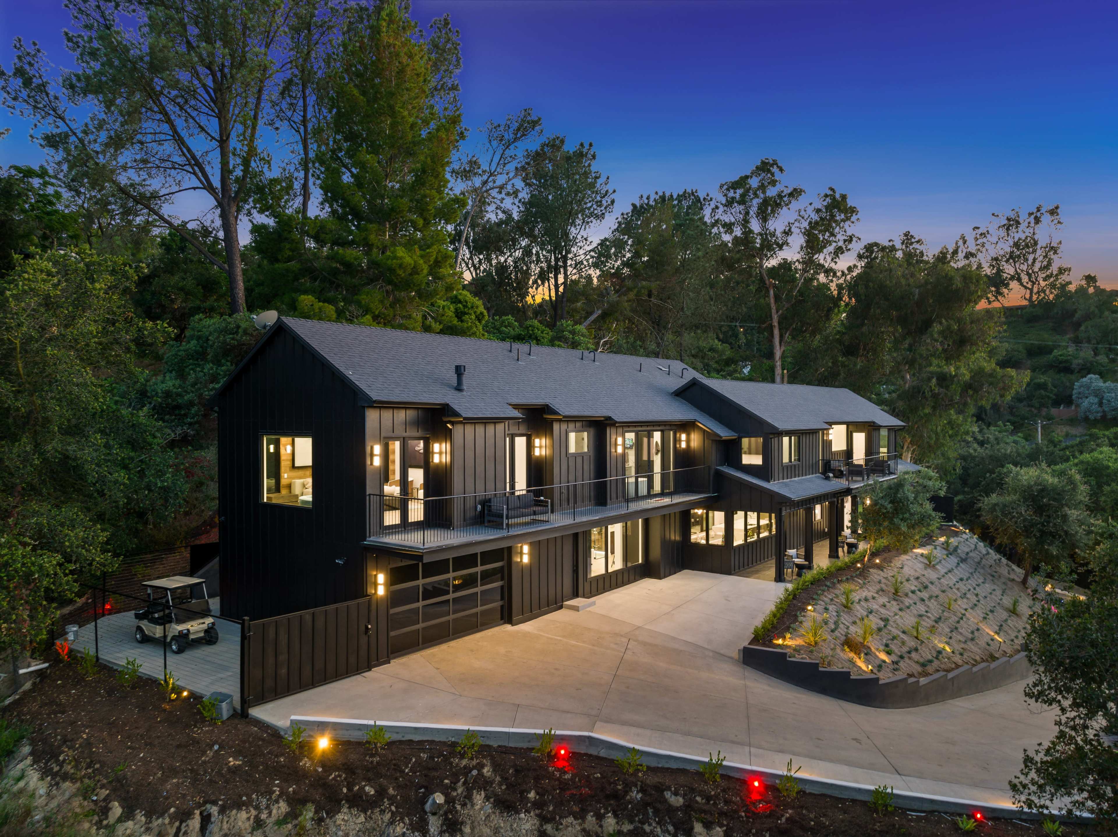 A modern black-clad house with multiple levels is situated on a landscaped hillside surrounded by trees at dusk.
