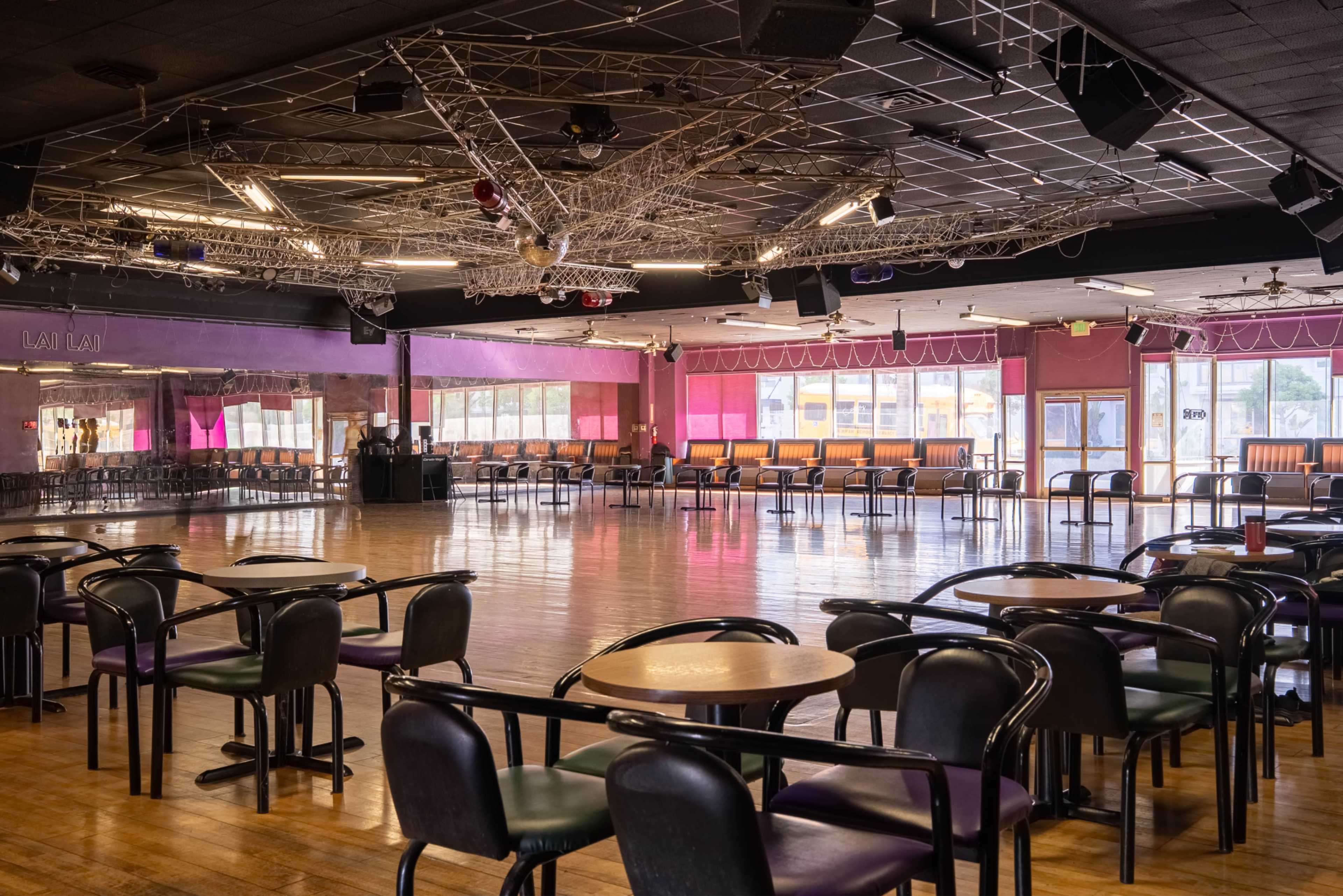 The image shows an empty dance hall with wooden floors, tables, and chairs arranged around the space, and a stage in the background.