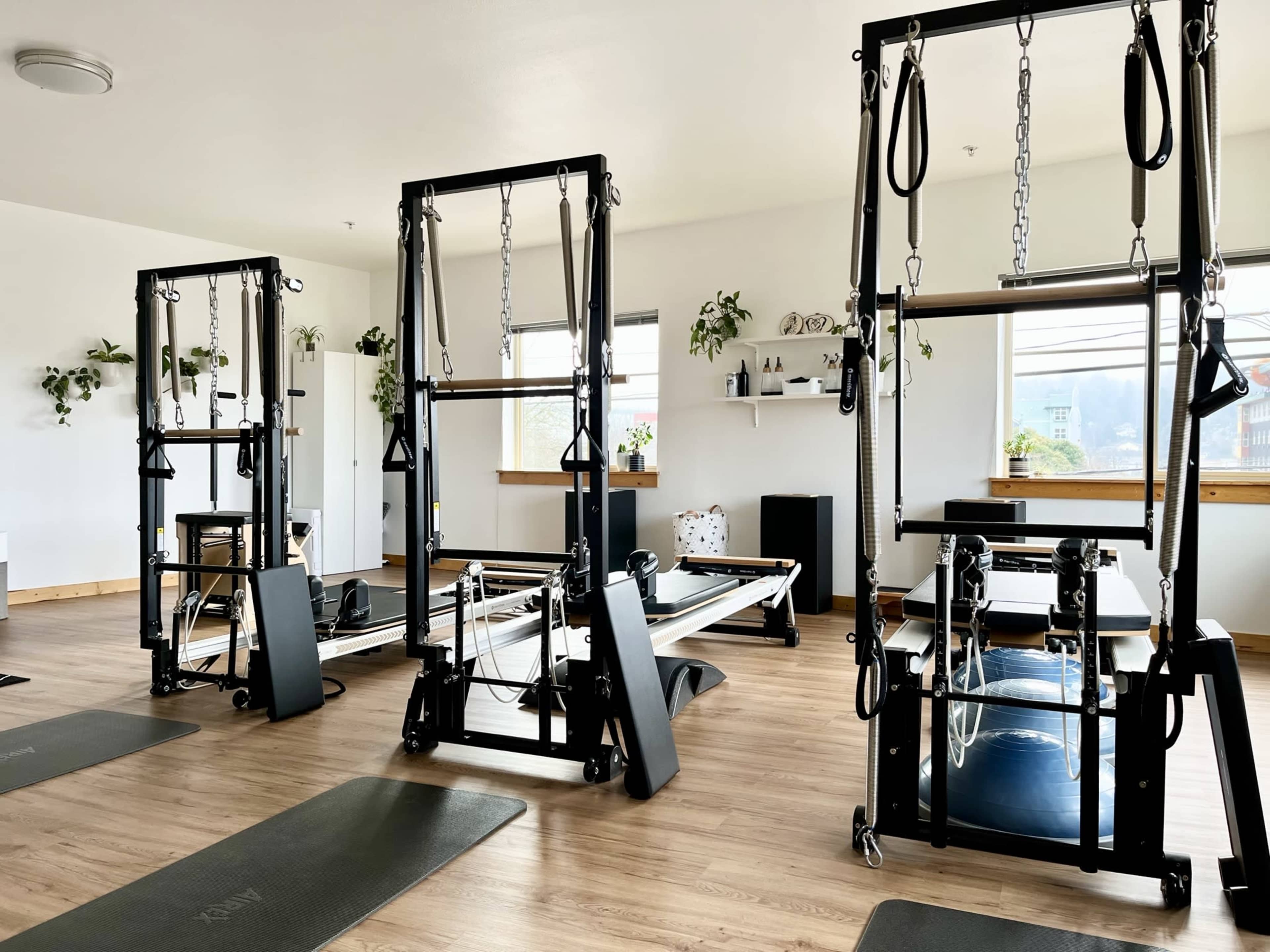A spacious and bright workout studio equipped with multiple reformer machines and neatly arranged exercise mats on a wooden floor.