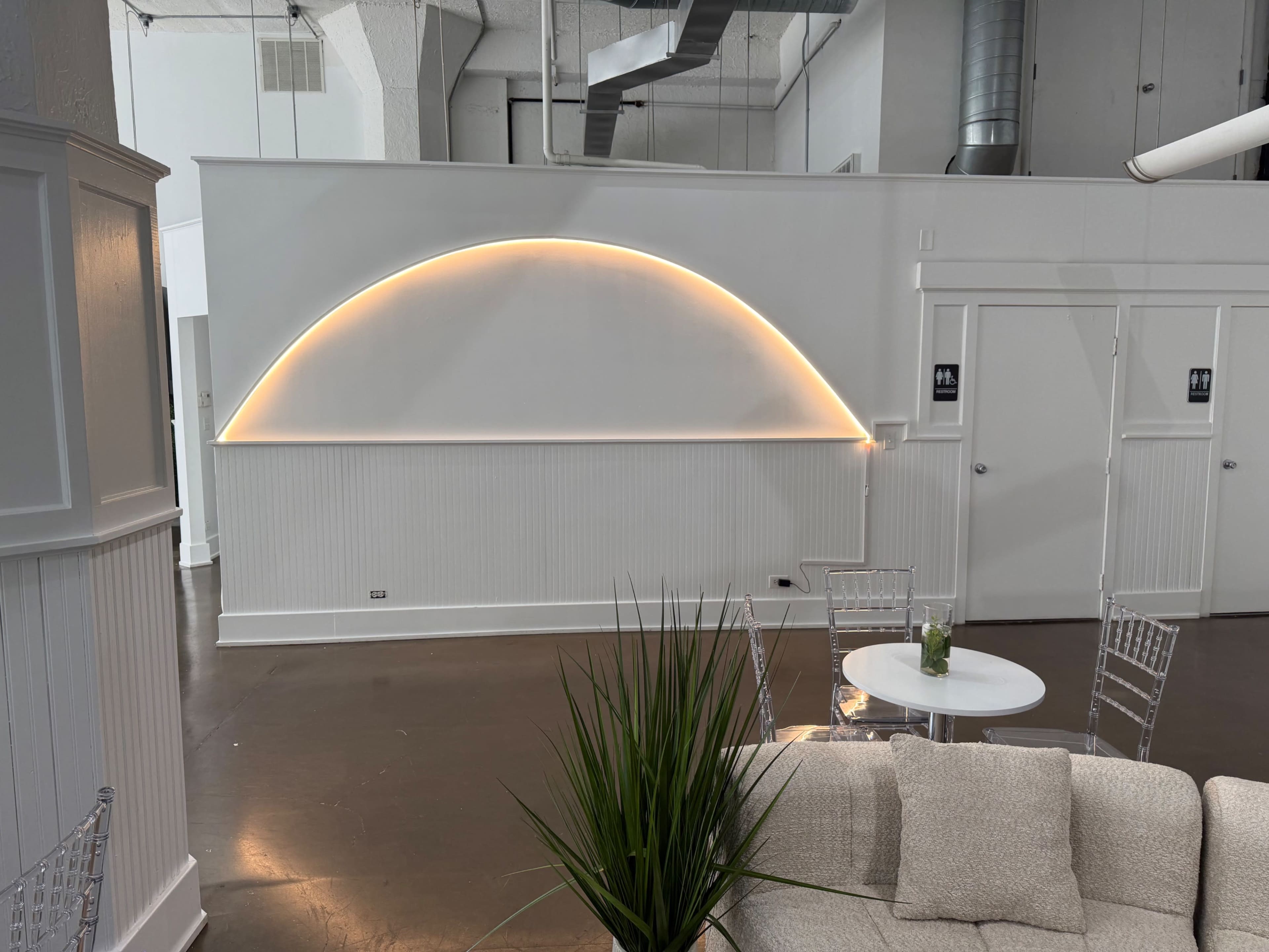The image shows a modern interior space featuring a white wall with a curved light fixture, a small round table, and clear chairs, complemented by a potted plant.