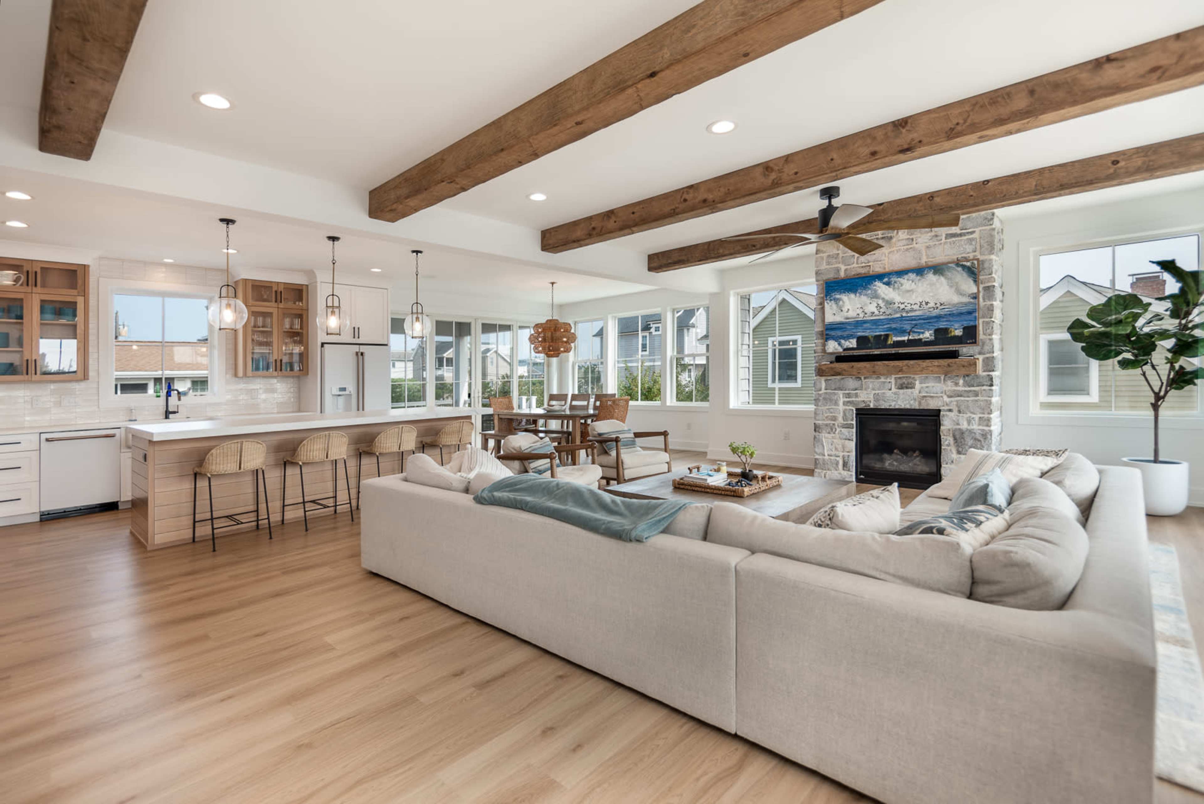 A spacious living area features a sectional sofa, wooden beams on the ceiling, and a stone fireplace, with a dining table and kitchen visible in the background.