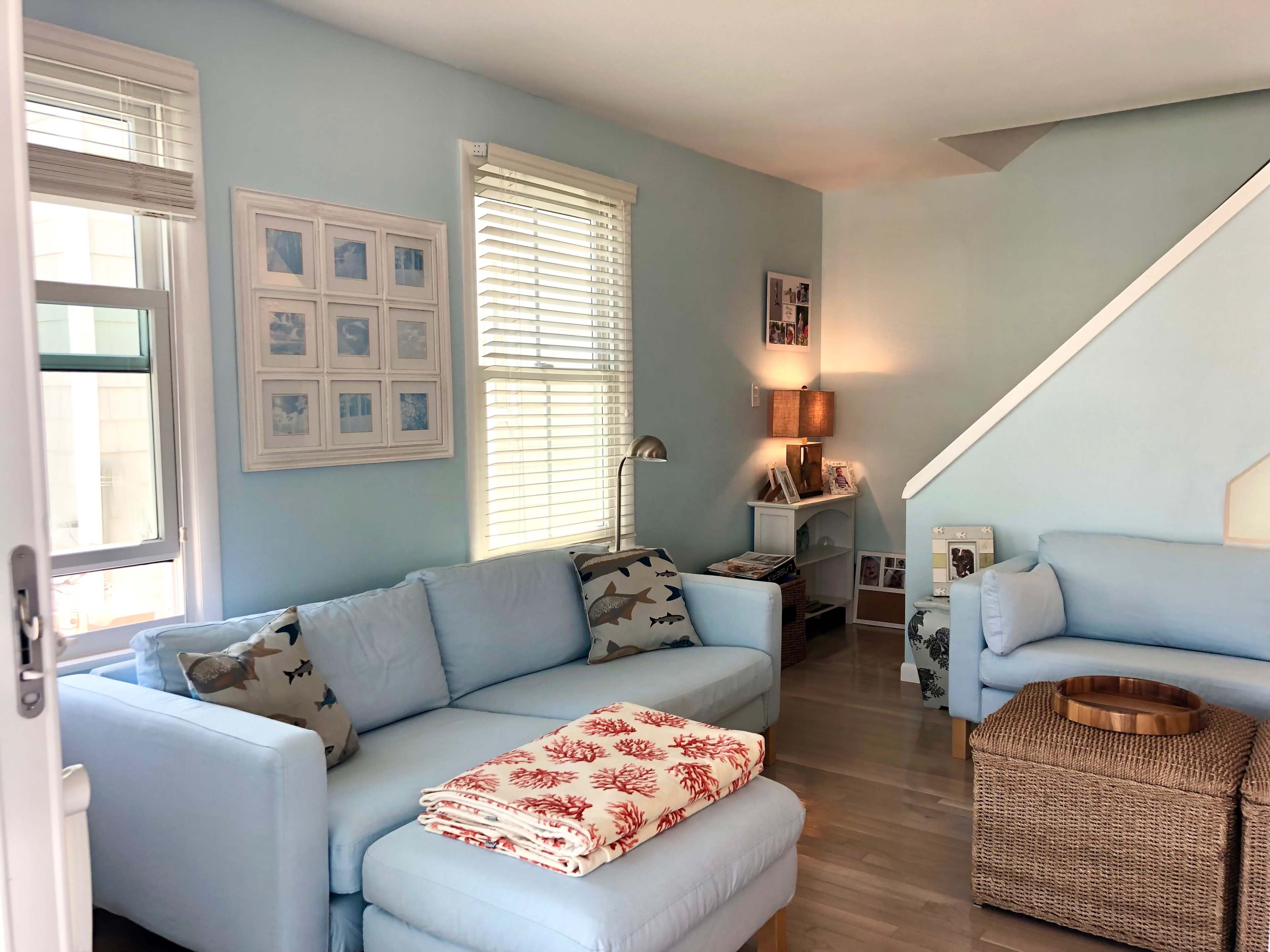 A light blue living room features a sectional sofa with coral-patterned throws, a woven coffee table, and framed photos on the walls.