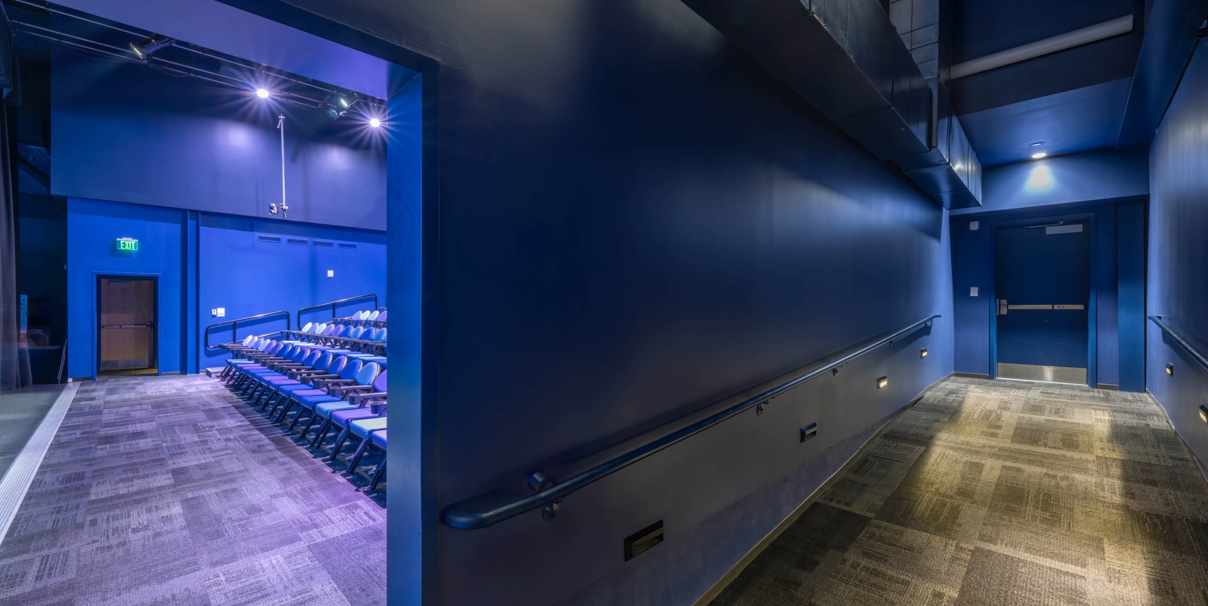 The image shows a dimly lit theater hallway leading to a set of blue walls and an exit door, with rows of empty seats visible in the background.