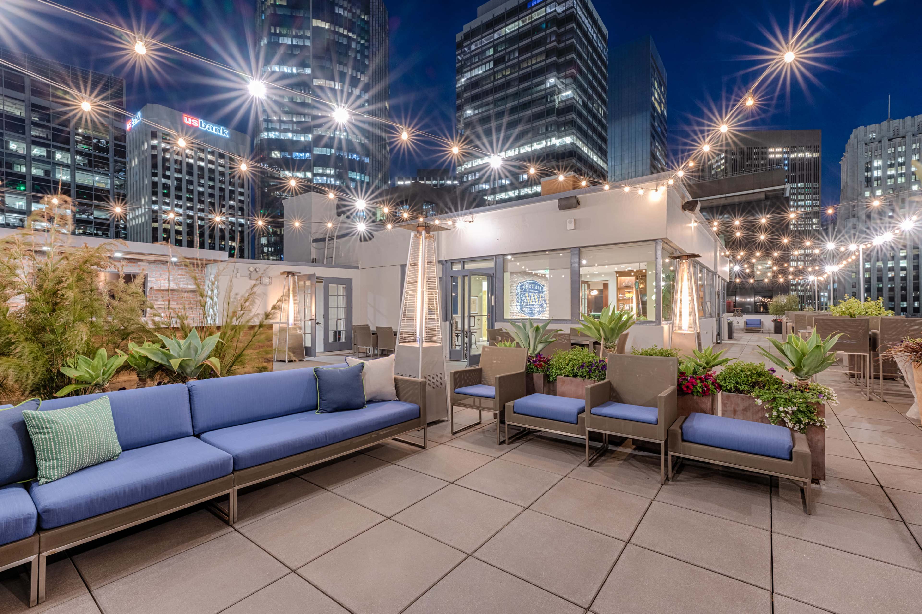 A rooftop lounge area features blue seating, potted plants, and string lights against a backdrop of city skyscrapers at night.