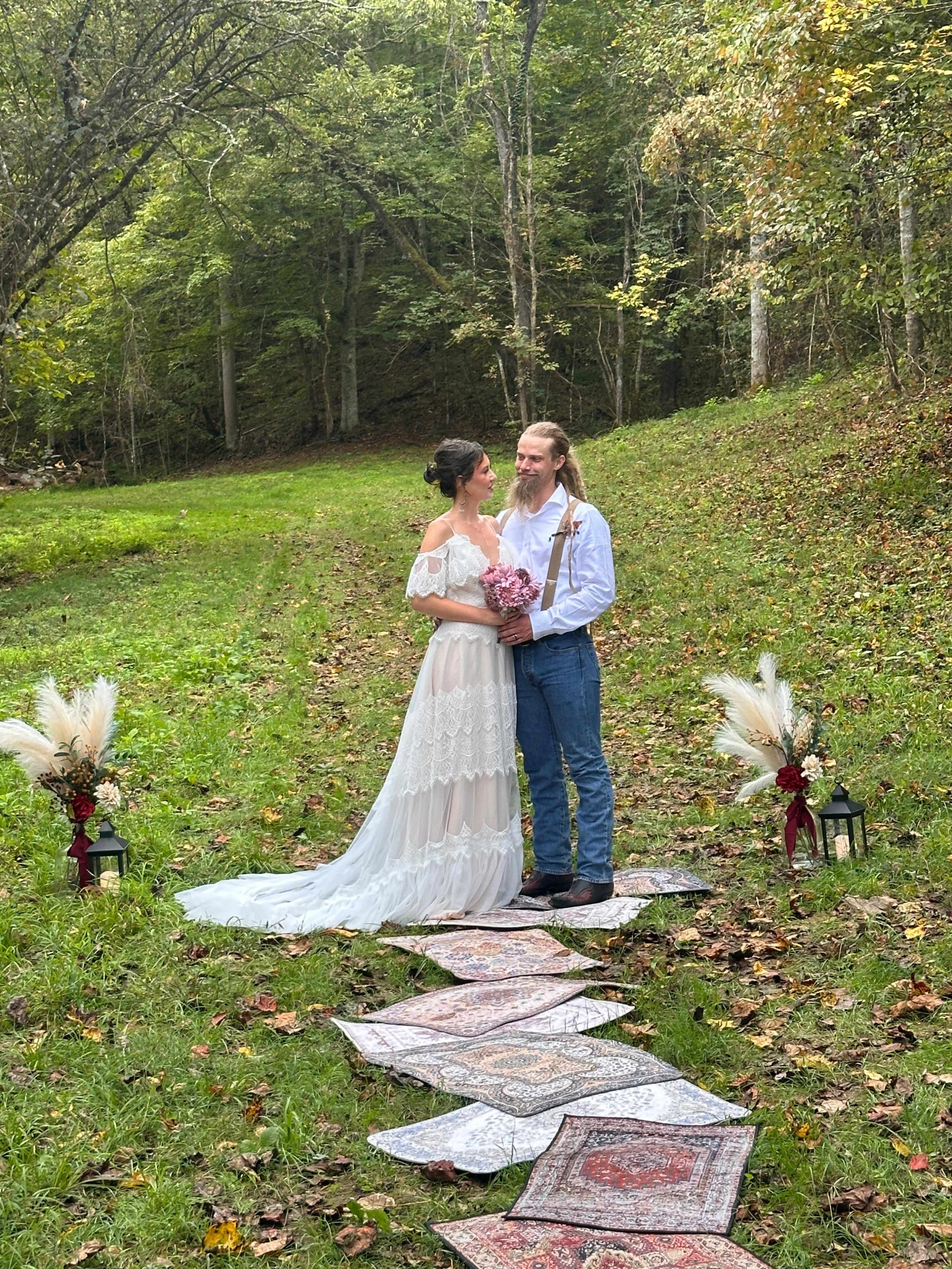 A couple stands together on a path lined with decorative rugs and lanterns, surrounded by trees and greenery in a natural setting.