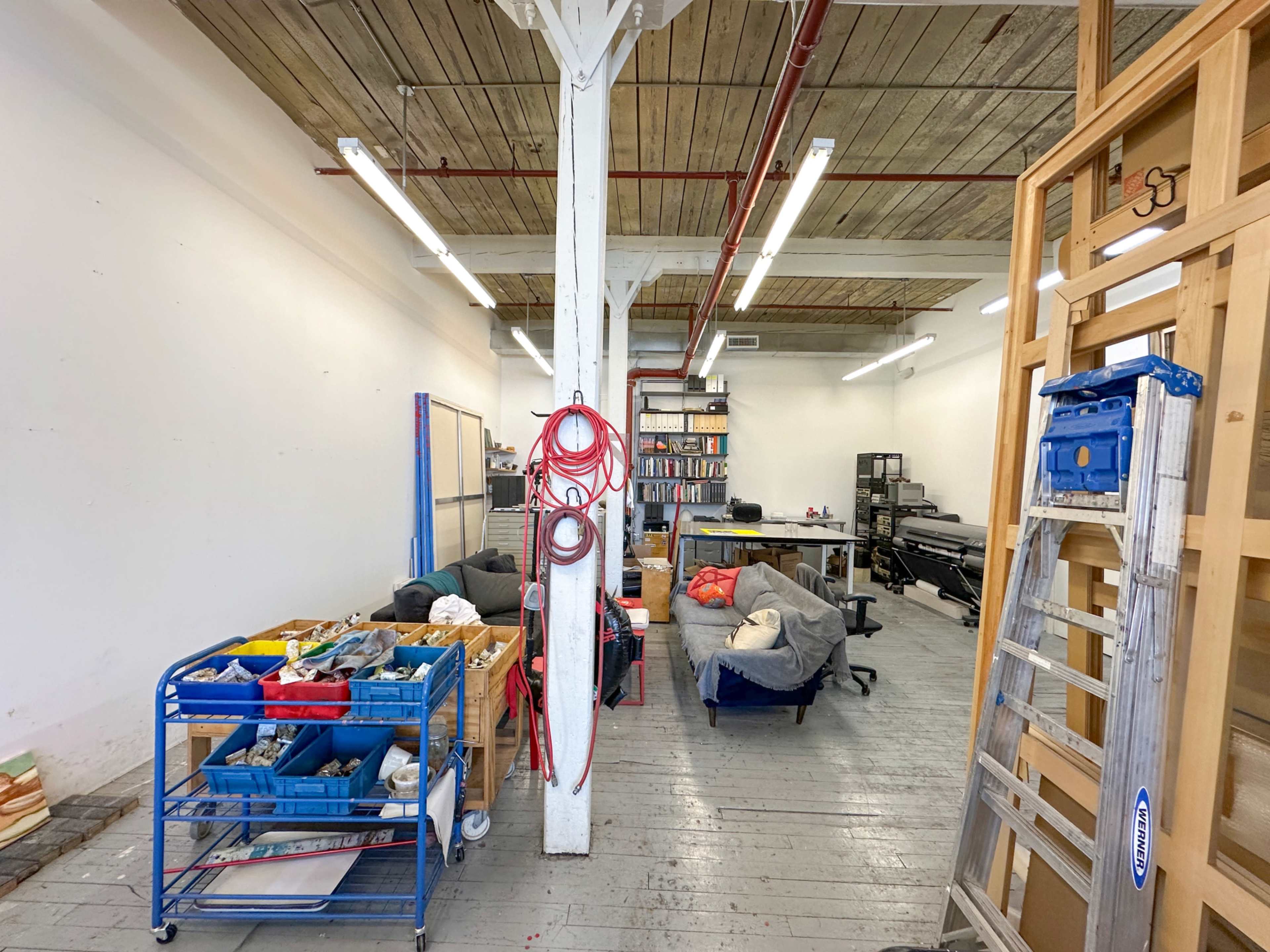 The image shows a large, open workspace with wooden flooring, shelves stacked with items, and various furniture pieces, including work tables and bean bags.