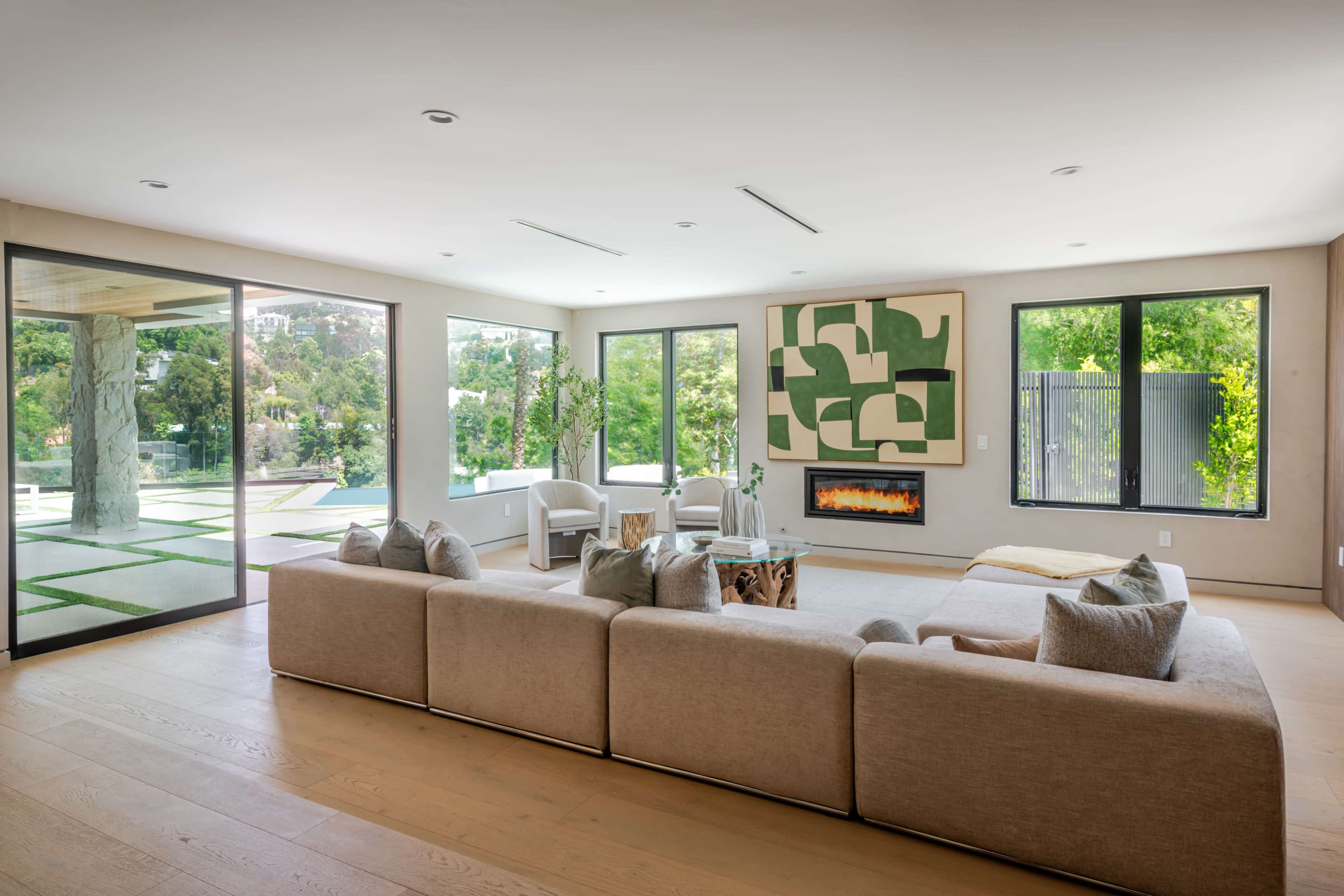 A modern living room features a large beige sectional sofa, abstract wall art, and expansive windows overlooking a landscaped outdoor area.
