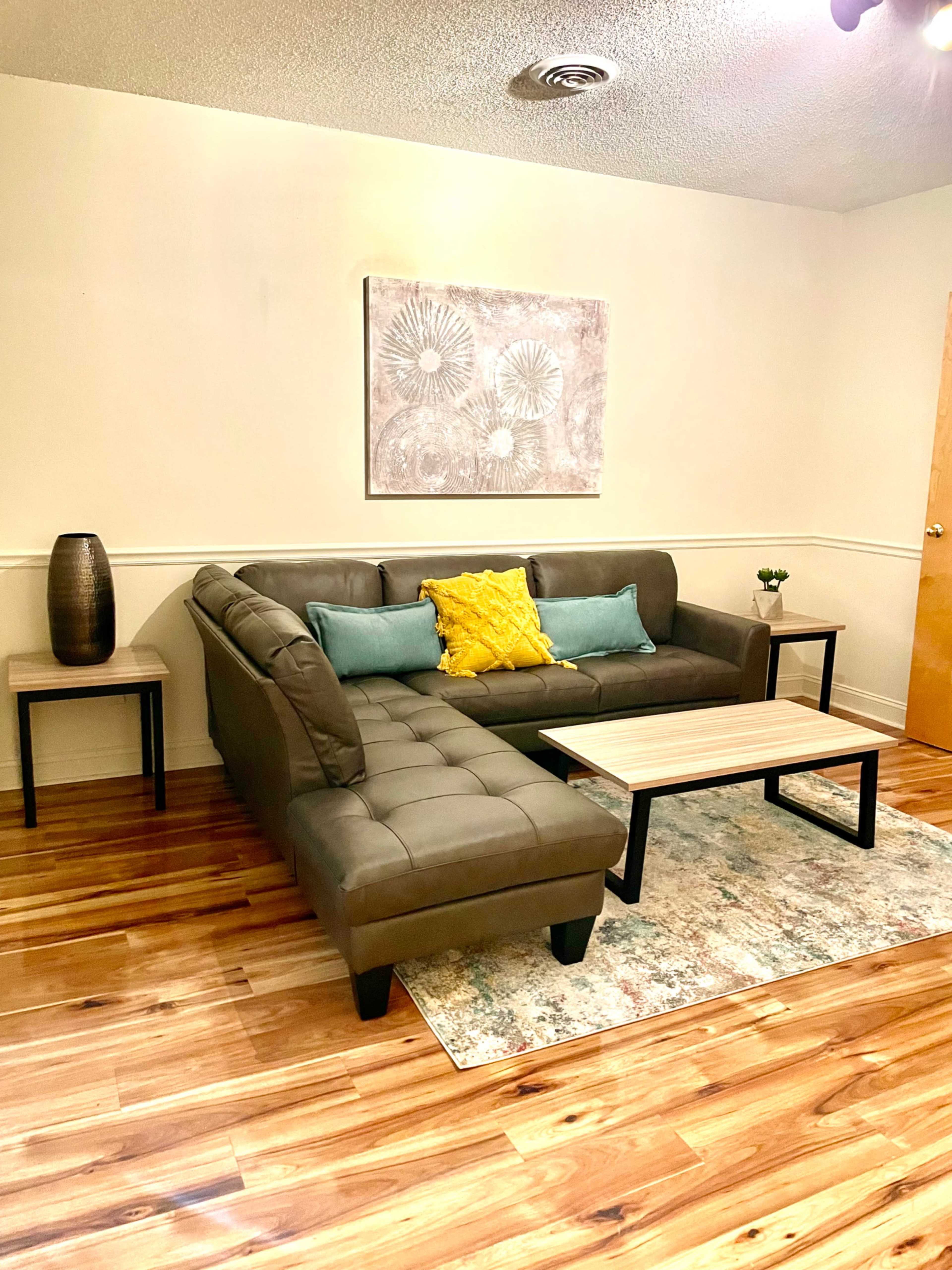 A modern living room features a gray sectional sofa with colorful cushions, a small wooden coffee table, and a decorative vase on a side table, all set on a wooden floor.