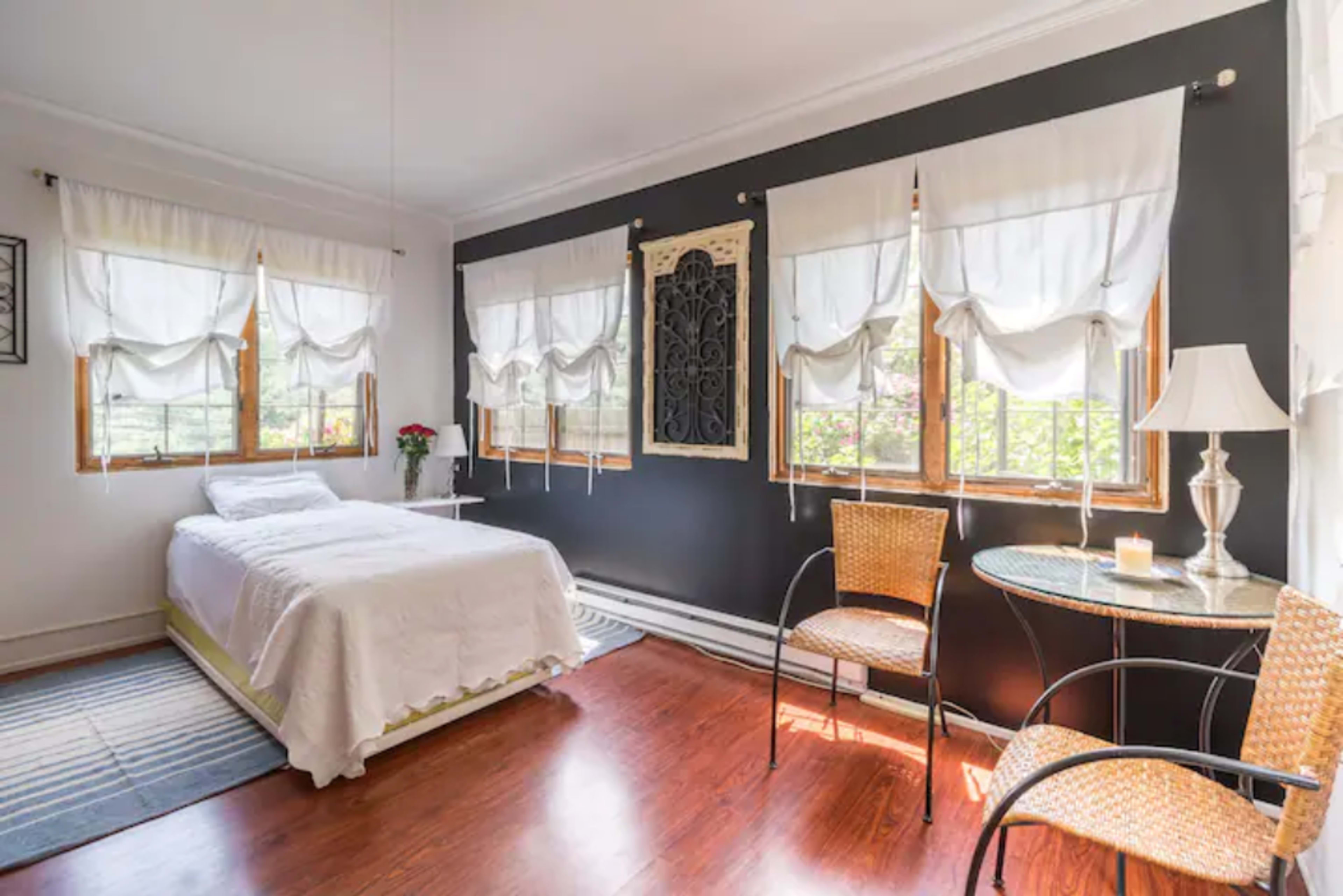 A small bedroom with a bed, two wicker chairs, a side table, and large windows adorned with white curtains.