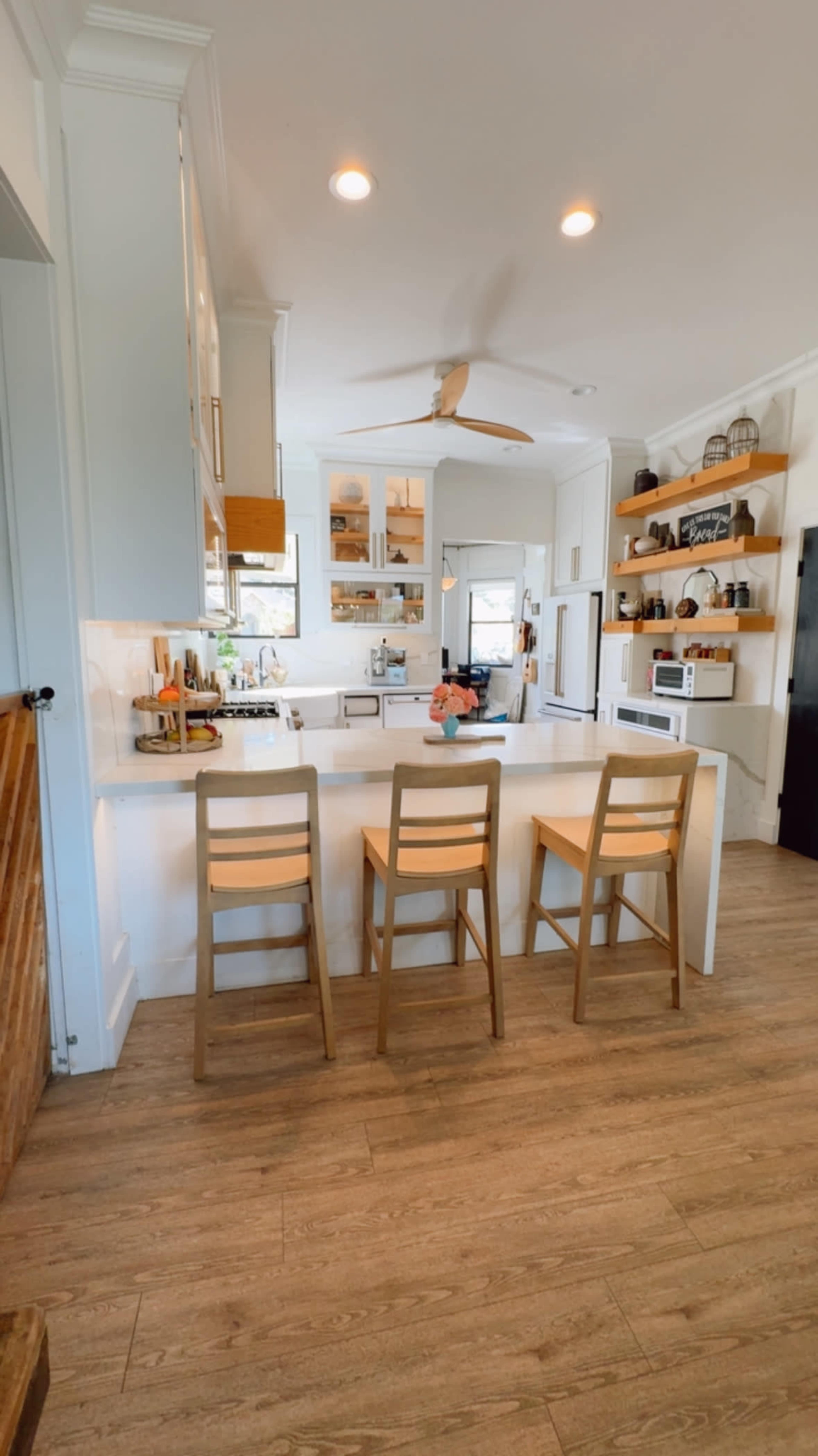 The image shows a modern kitchen featuring a central island with three wooden bar stools, light cabinetry, open shelving, and a ceiling fan.