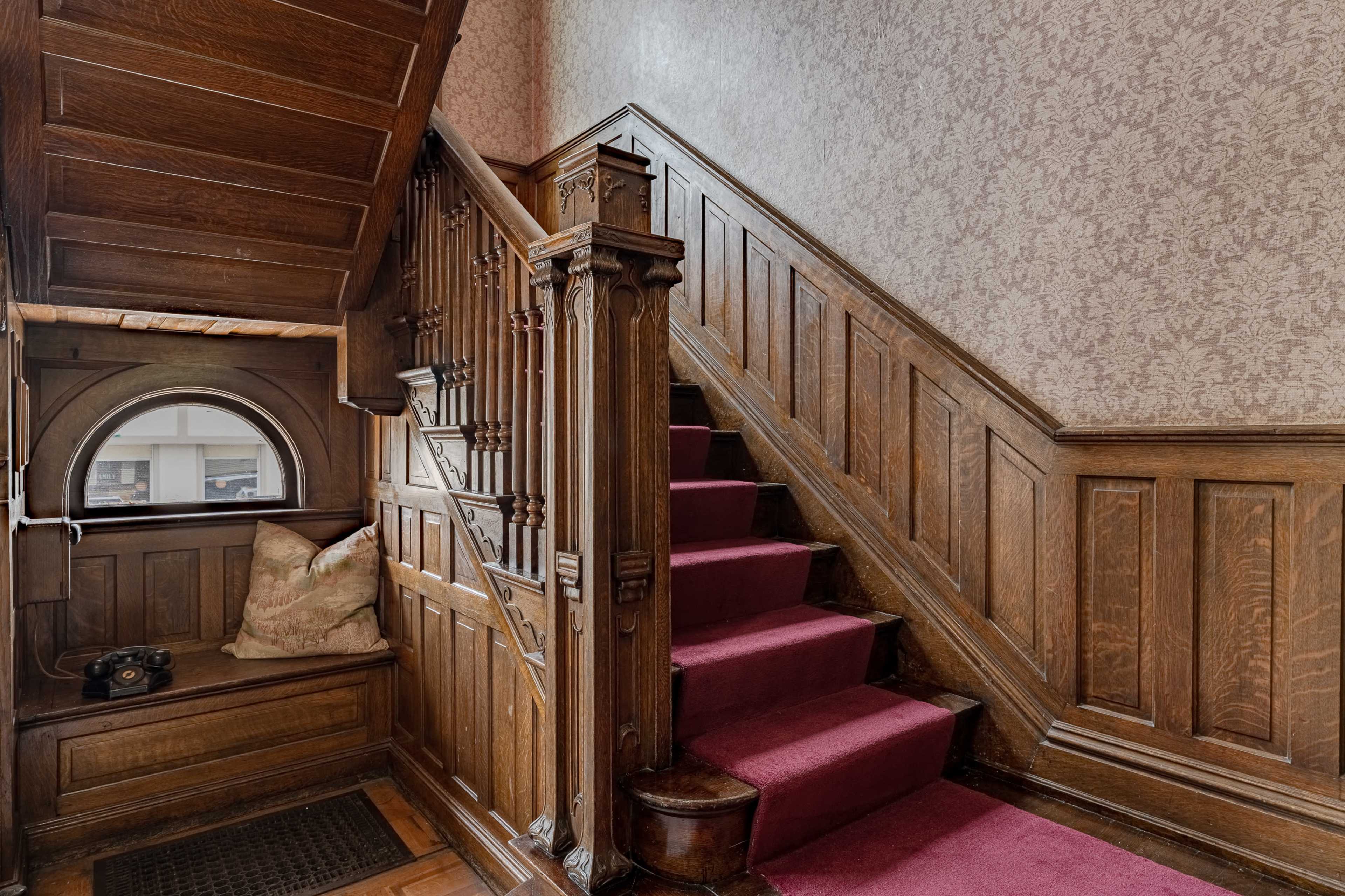 A wooden staircase with a burgundy carpet leading to an upper floor, surrounded by intricately designed wood paneling and a decorative window nook.