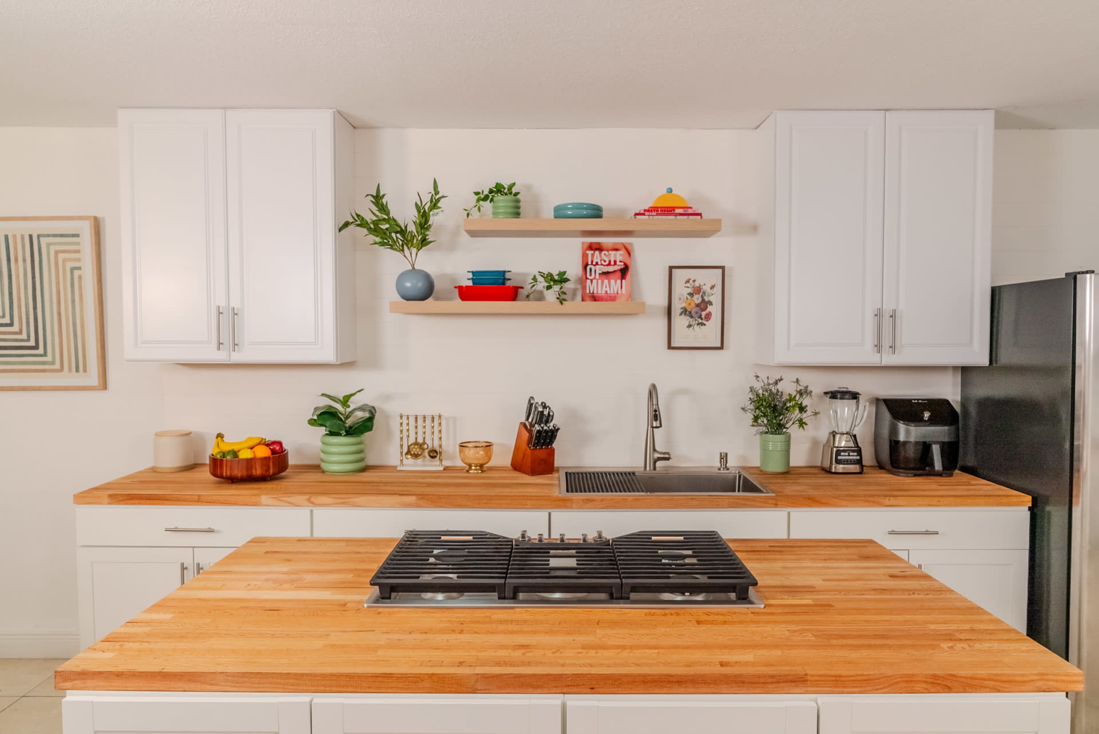 A modern kitchen features a central wooden island with a gas stove, white cabinetry, and open shelves displaying decorative items and plants.