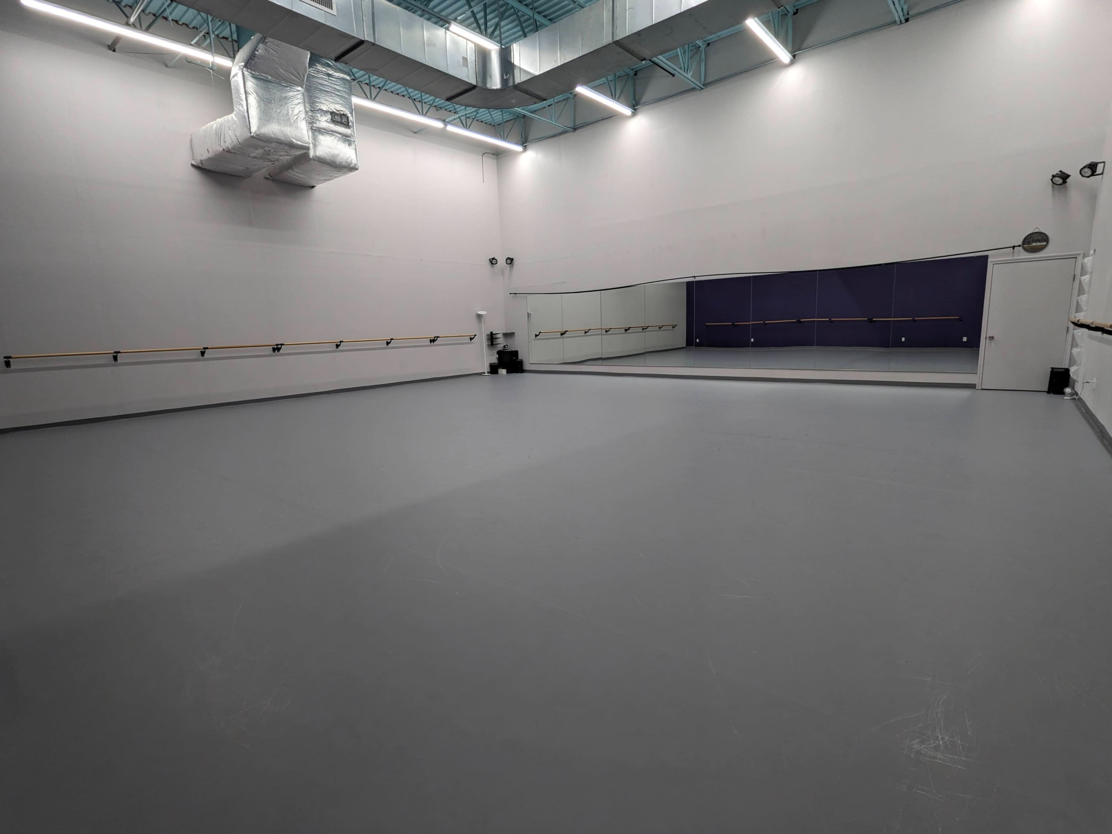 The image shows a spacious dance studio with a gray floor, mirrors on one wall, and barre along the sides.