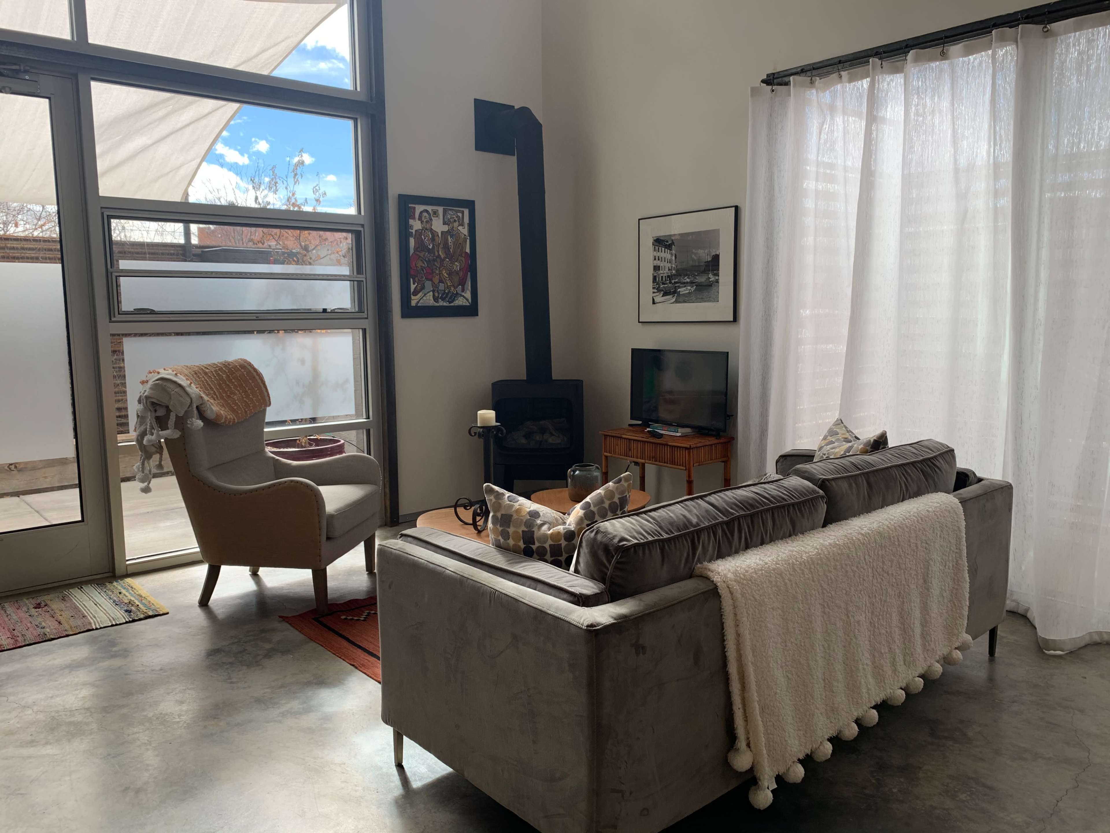 A modern living room features a gray sofa with a white throw blanket, a television on a wooden stand, and a cozy armchair near a glass door leading to an outdoor space.