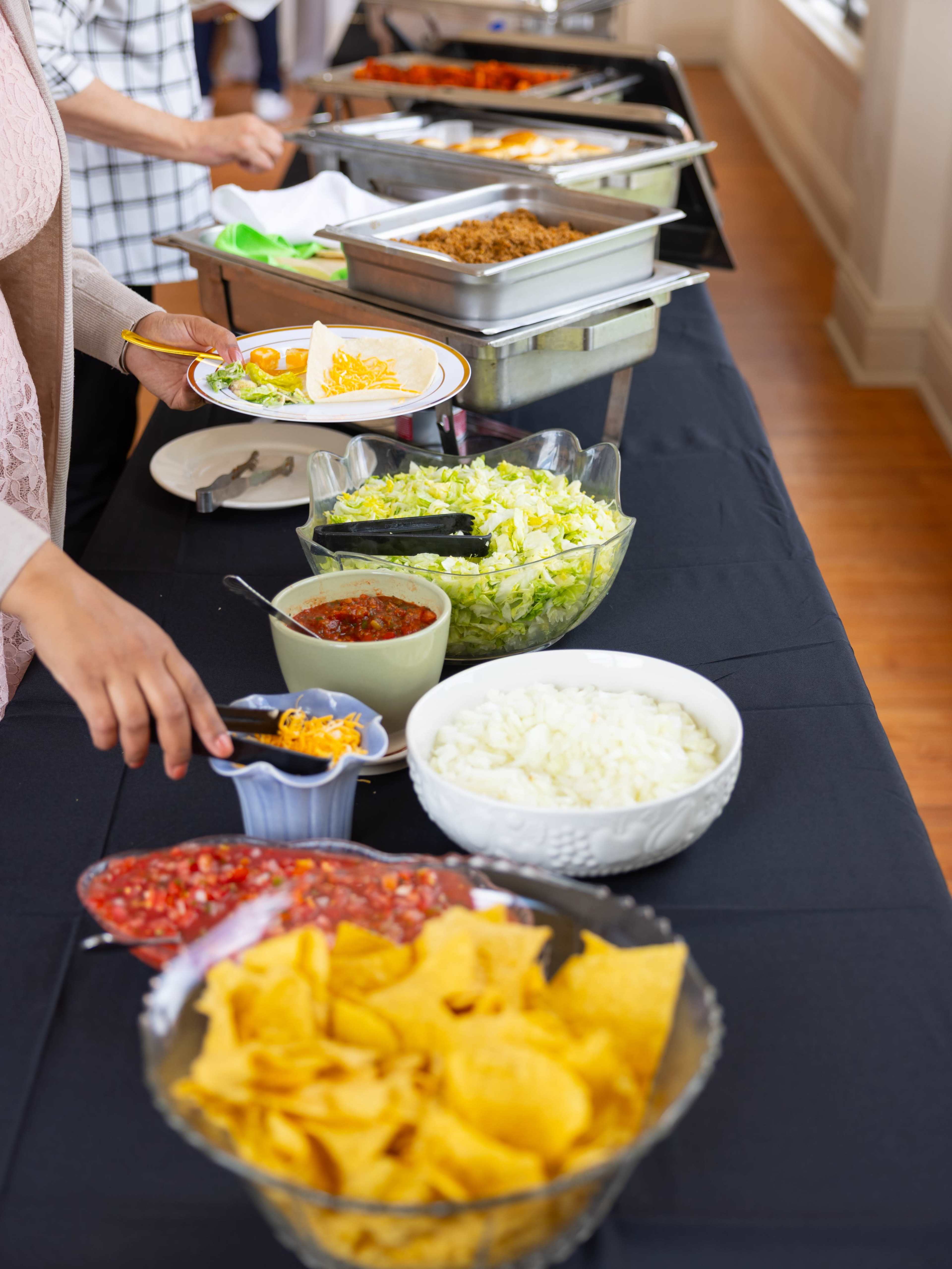 A person is serving food at a buffet table featuring various toppings and ingredients for tacos, including lettuce, cheese, salsa, and tortilla chips.