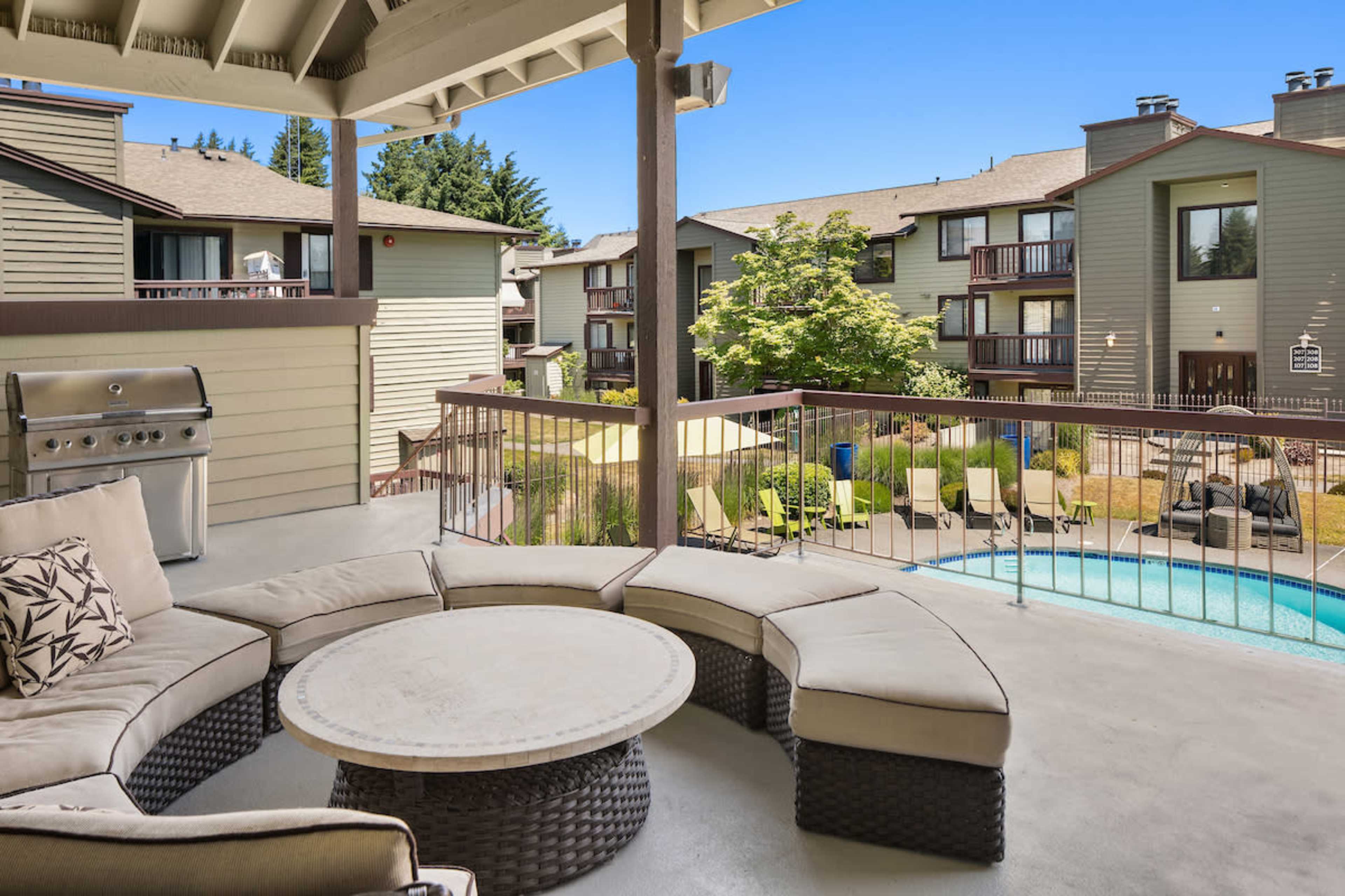 A patio with a circular seating area and a grill overlooks a pool surrounded by landscaped greenery and apartment buildings.