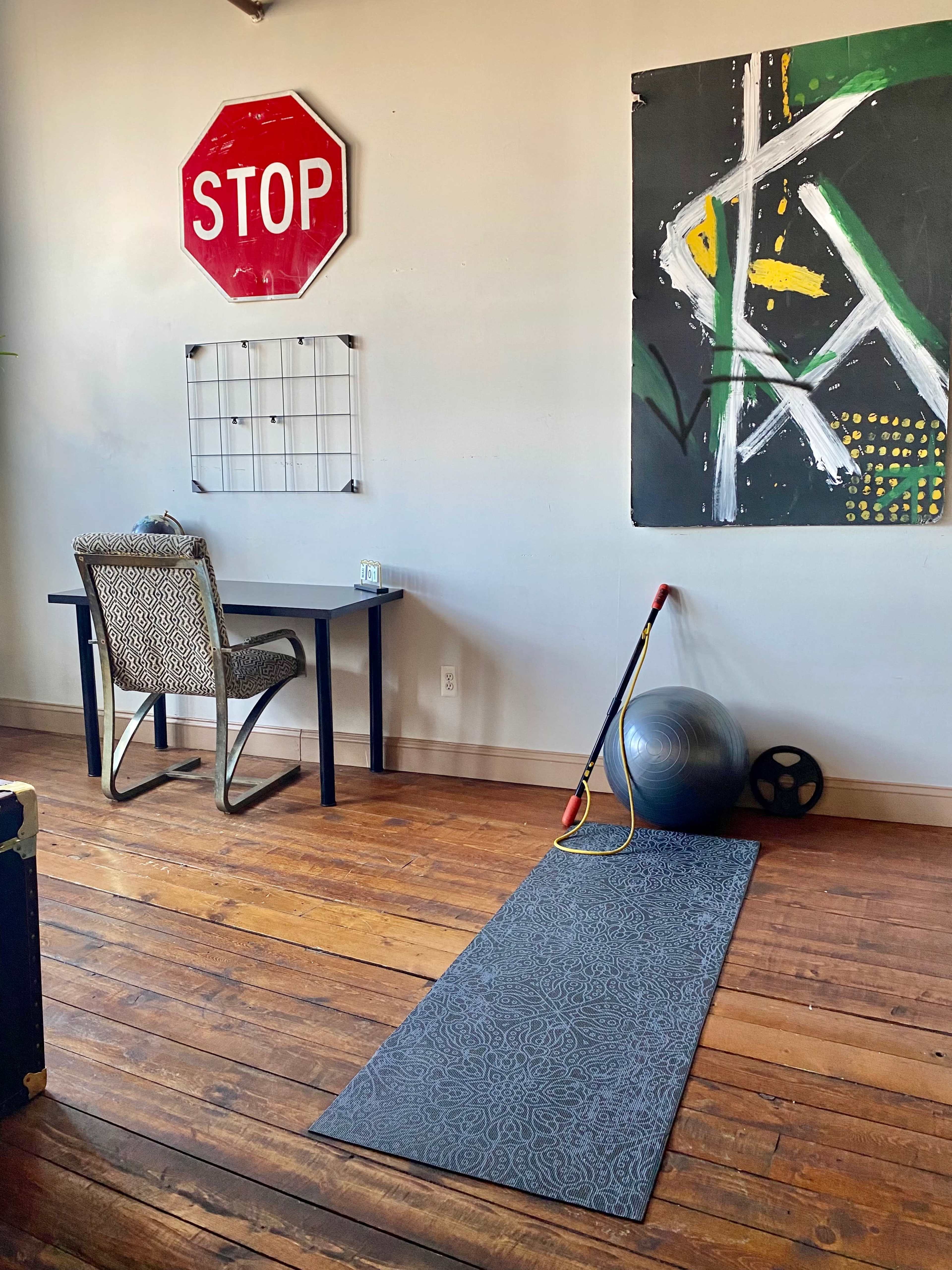 A room with a wooden floor featuring a desk, a yoga mat, fitness equipment, and a large red stop sign on the wall.