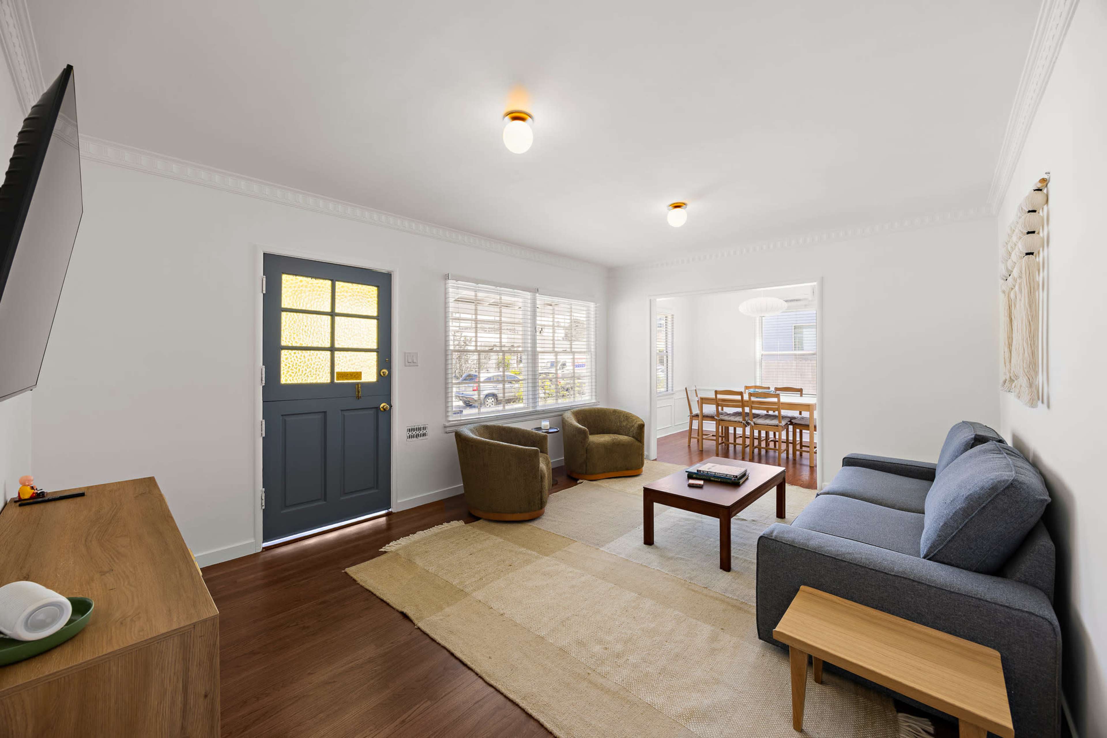 The image shows a bright living room with a blue sofa, a wooden coffee table, two curved armchairs, and a door leading to the outside.