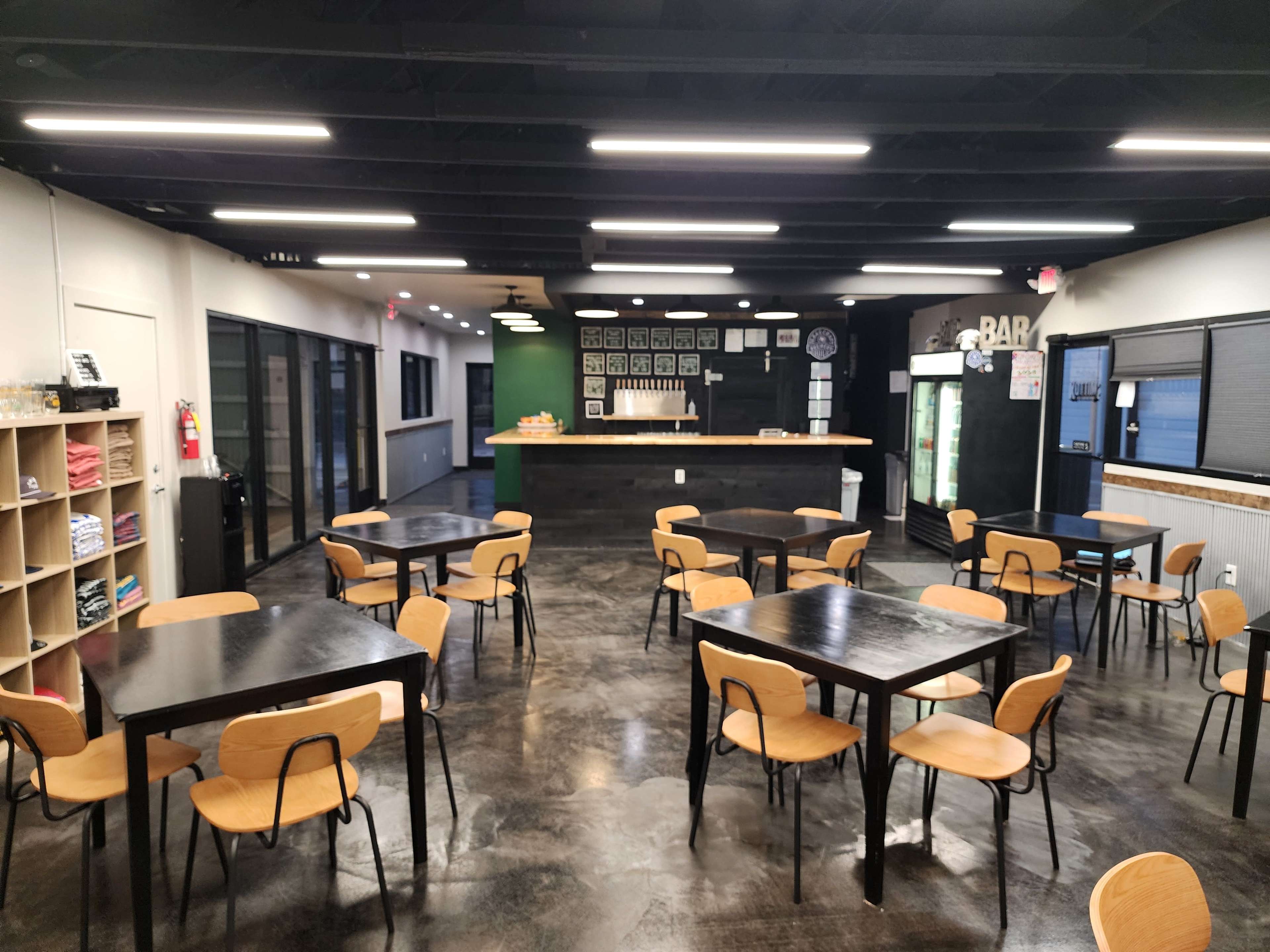 The image shows a spacious, modern interior of a café or bar, featuring several black tables with wooden chairs arranged neatly and a counter displaying various items in the background.