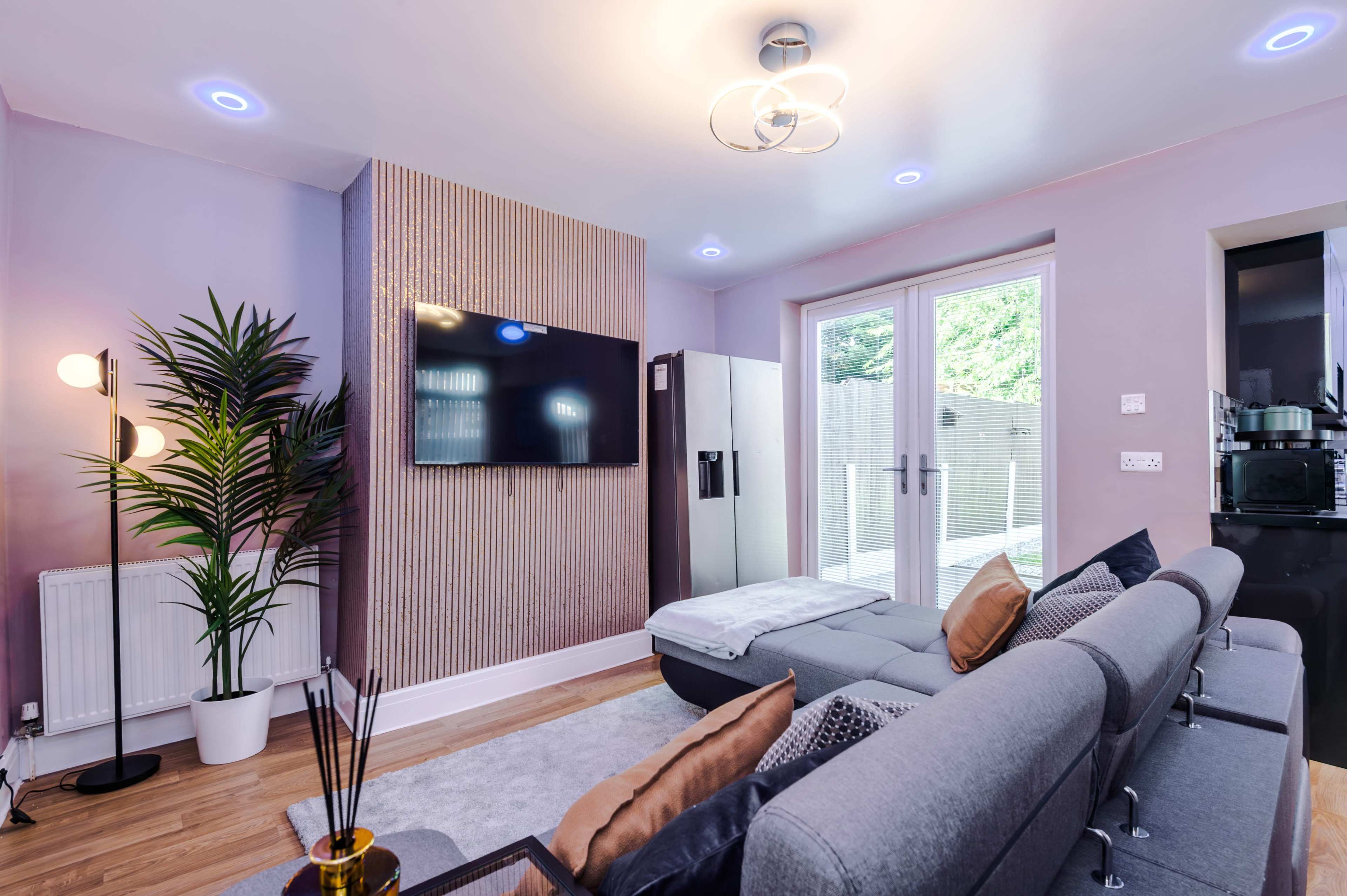 The image shows a modern living room with a gray sofa, a wall-mounted television, and a sliding glass door leading to an outdoor area, complemented by indoor plants and contemporary lighting.