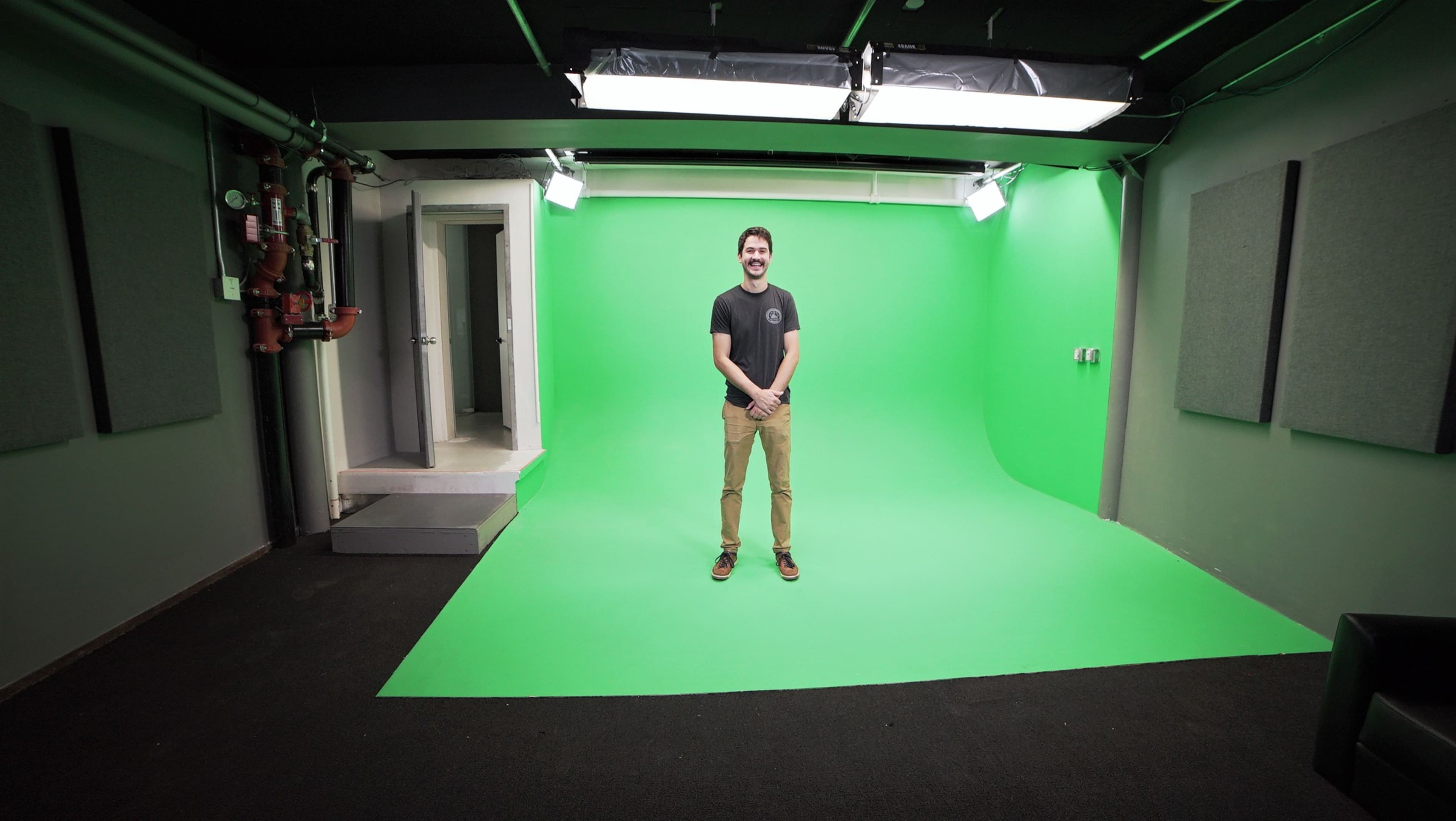 A man stands on a green backdrop in a studio space with gray walls and bright overhead lights.