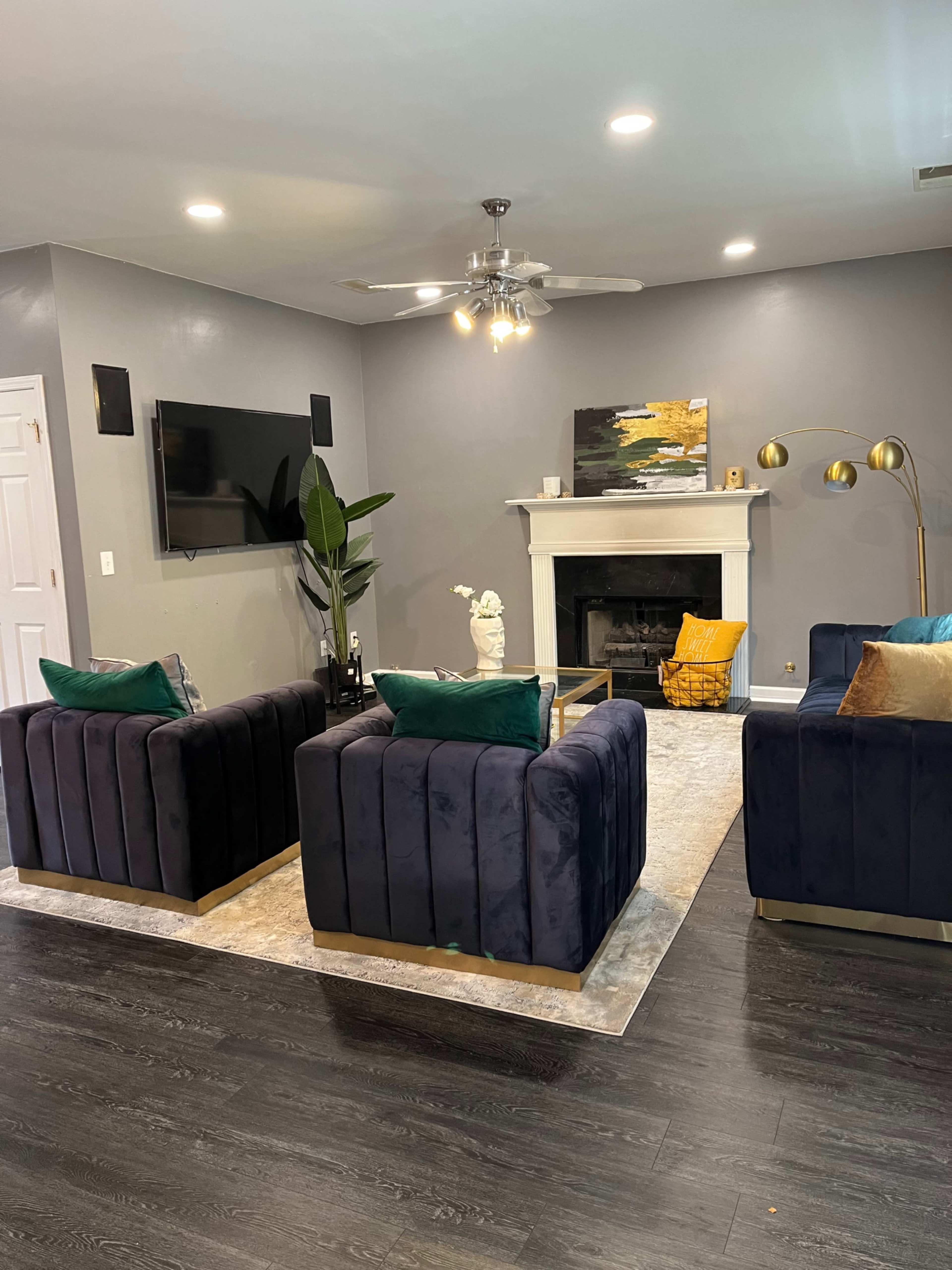 The living room features two velvet armchairs facing a modern fireplace, with a television mounted on the wall above and a decorative painting nearby.