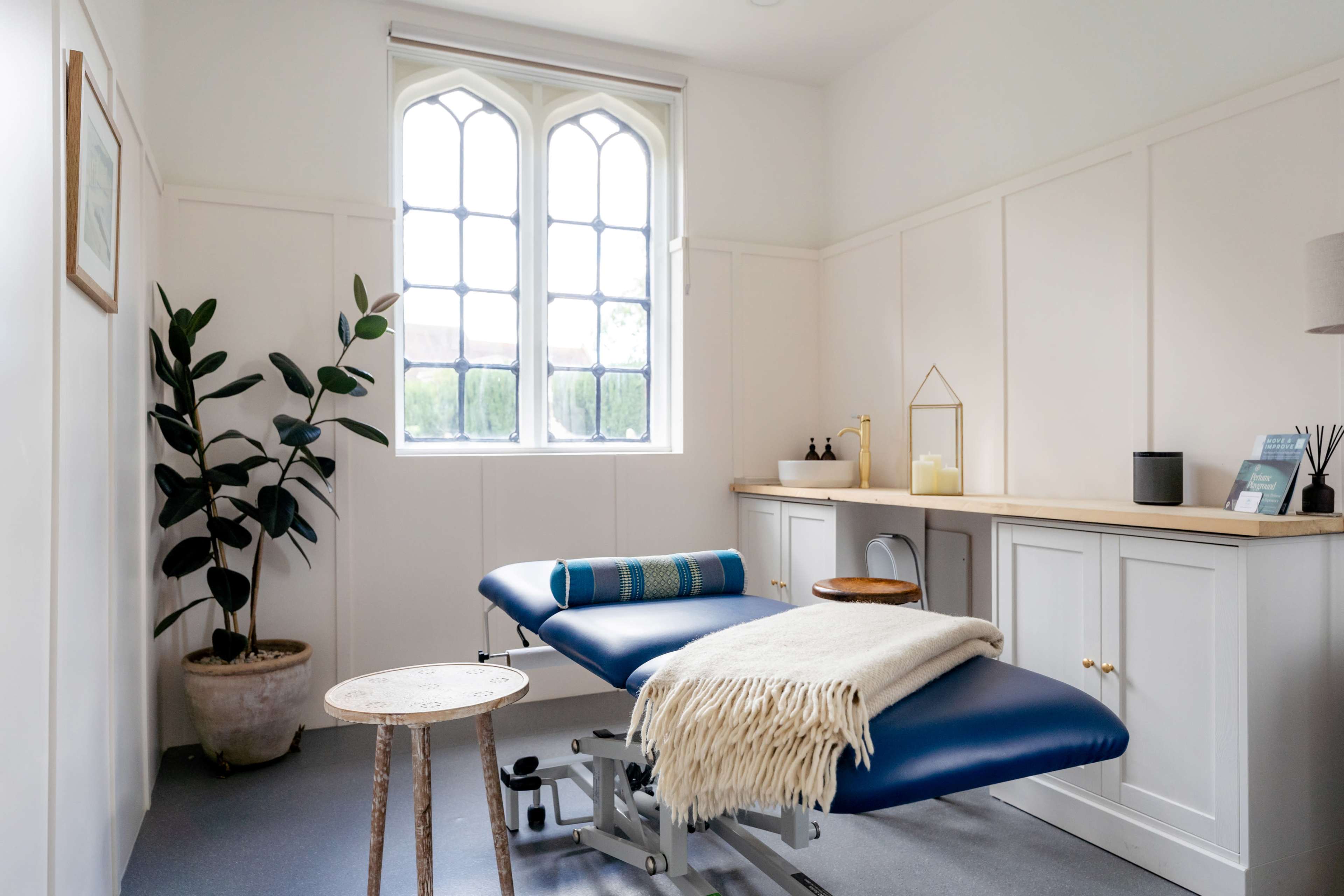 A minimalistic treatment room featuring a blue massage table, a small wooden table, a plant, and a large window with a decorative frame.