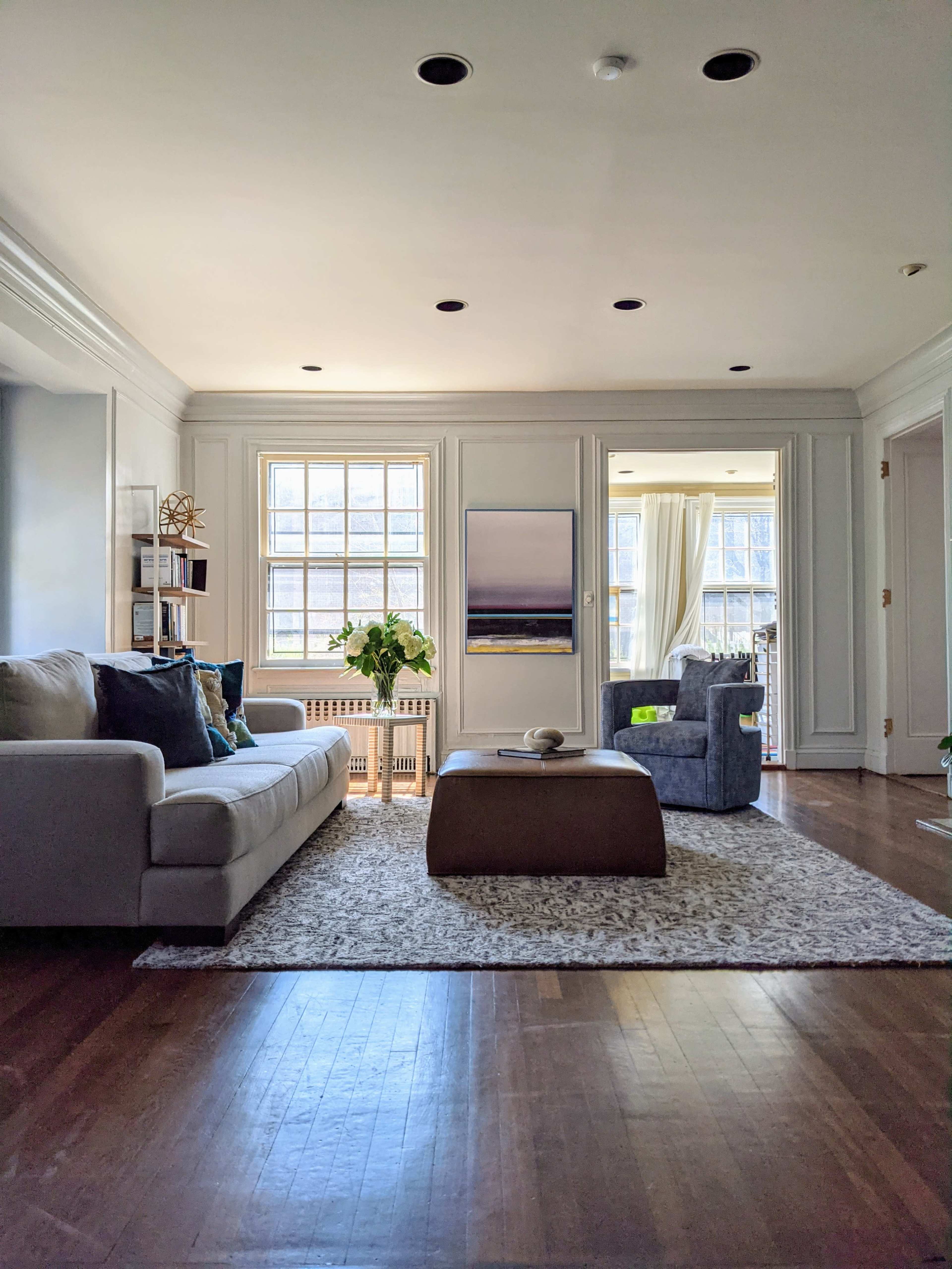 The living room features a light-colored sofa, a round coffee table, and a cozy armchair, with large windows providing ample natural light.