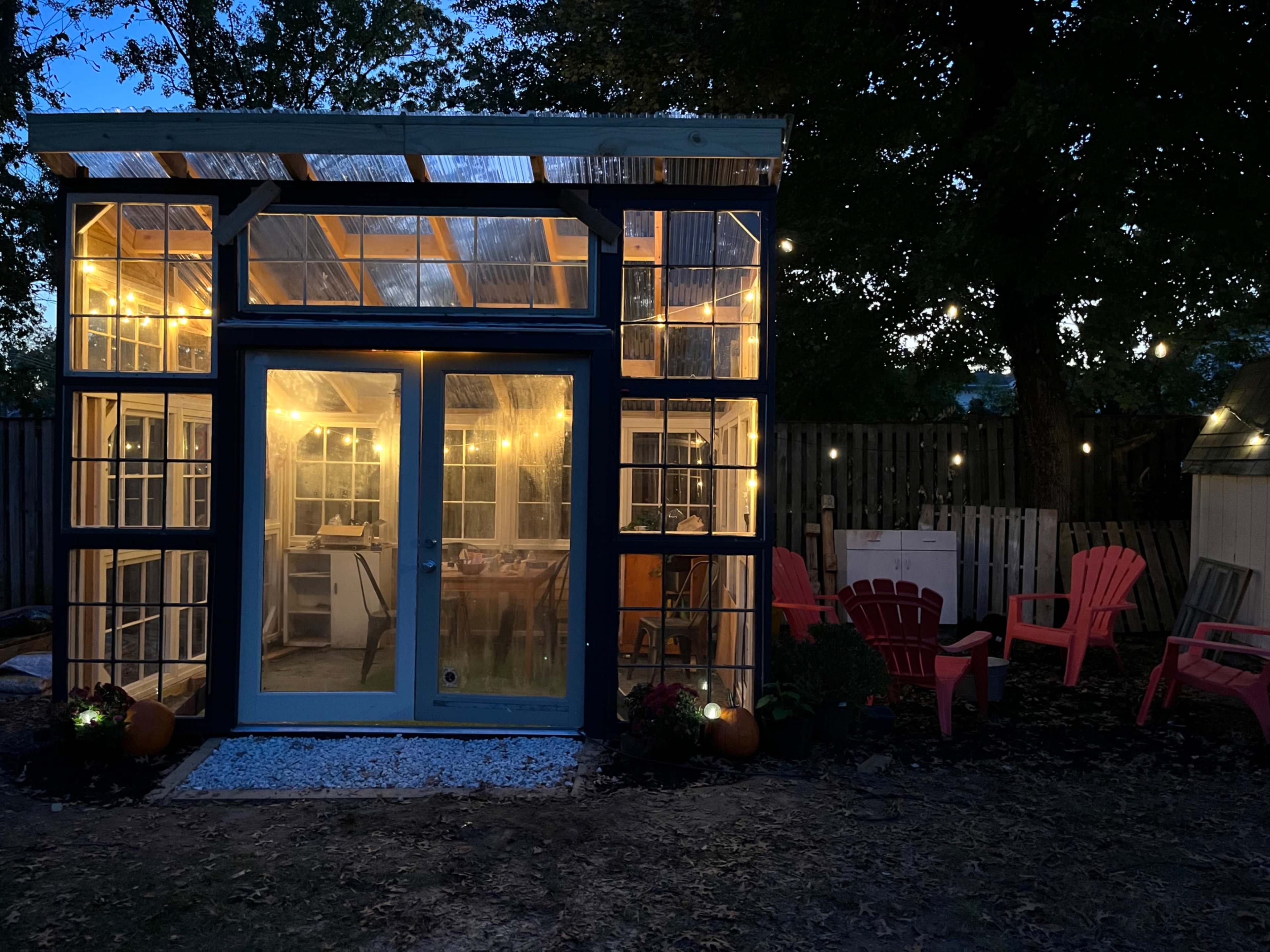 A glass-enclosed greenhouse with string lights and pink Adirondack chairs is illuminated at dusk.