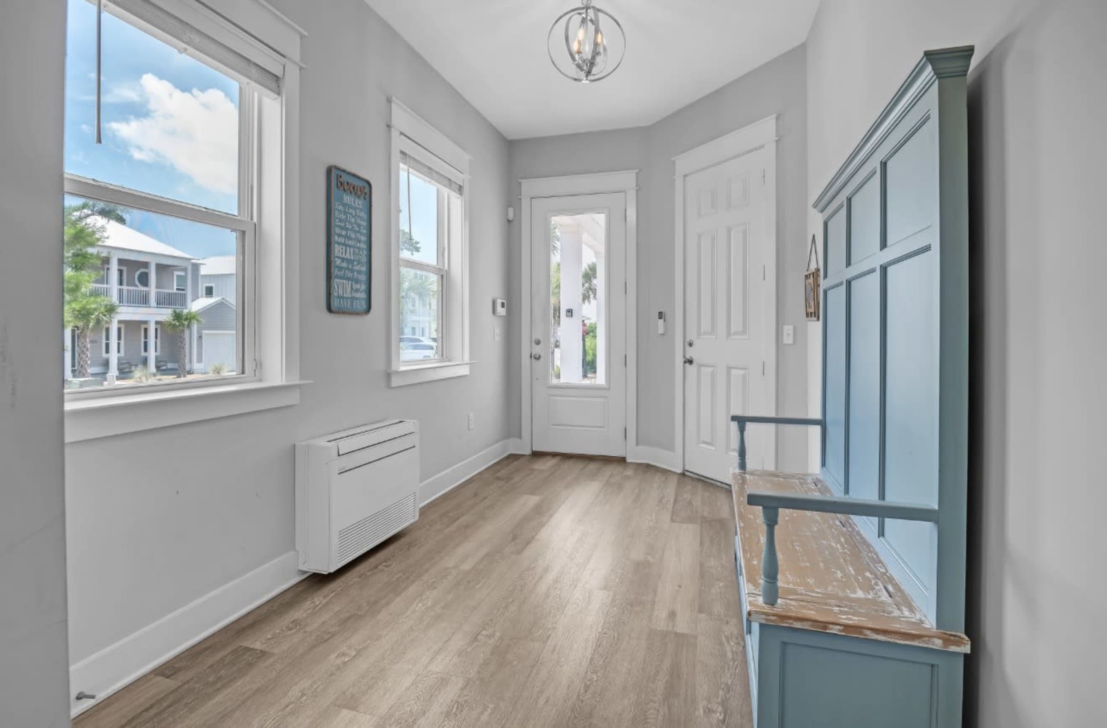 The image shows a spacious entryway with gray walls, a wooden floor, a front door, and a blue bench against the wall.