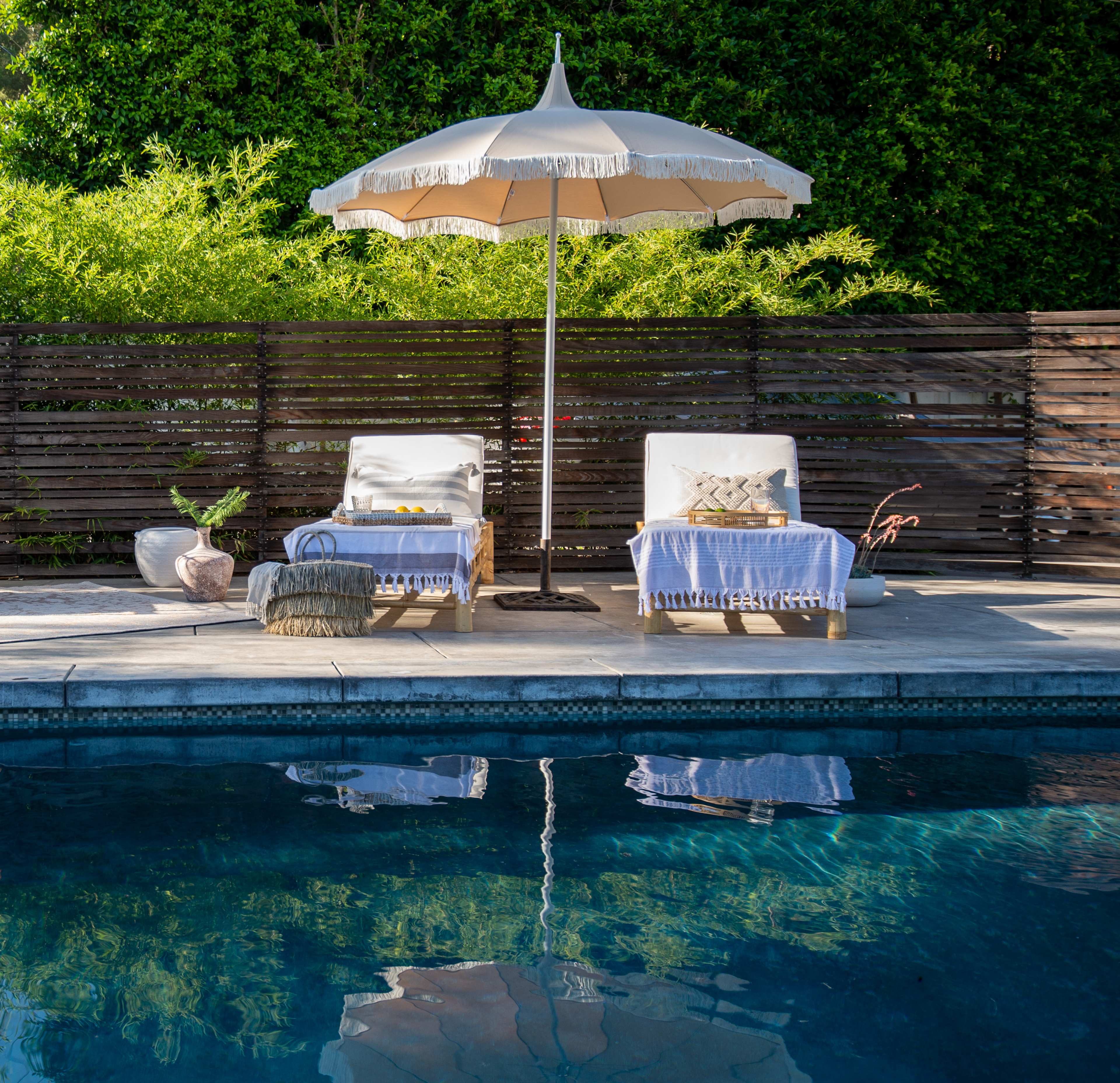 A pool area features two white lounge chairs with a large umbrella, surrounded by greenery and a wooden privacy screen.
