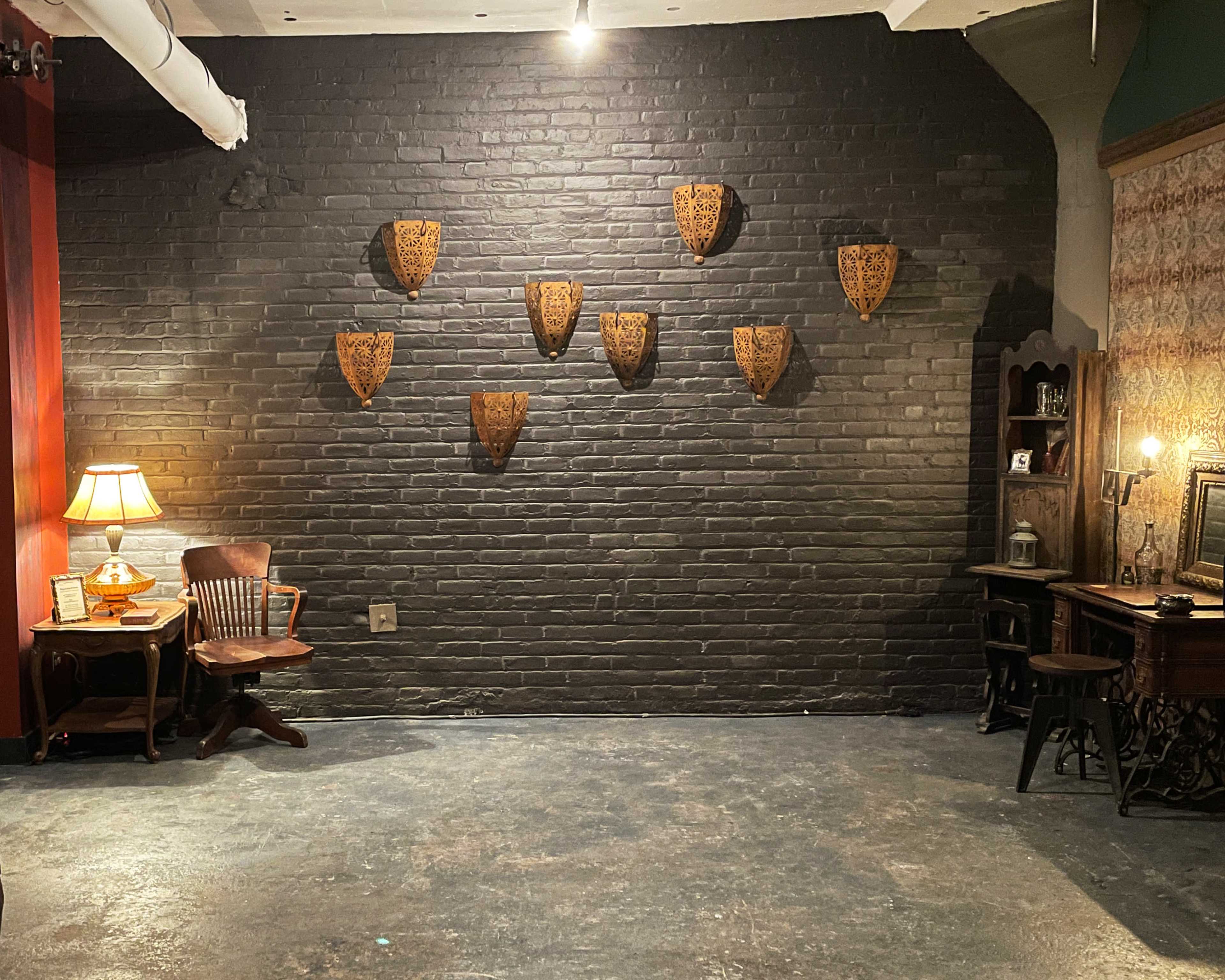 The image shows a dimly lit room with a black brick wall adorned with eight decorative wooden shields, a vintage lamp, and a wooden desk with a chair.