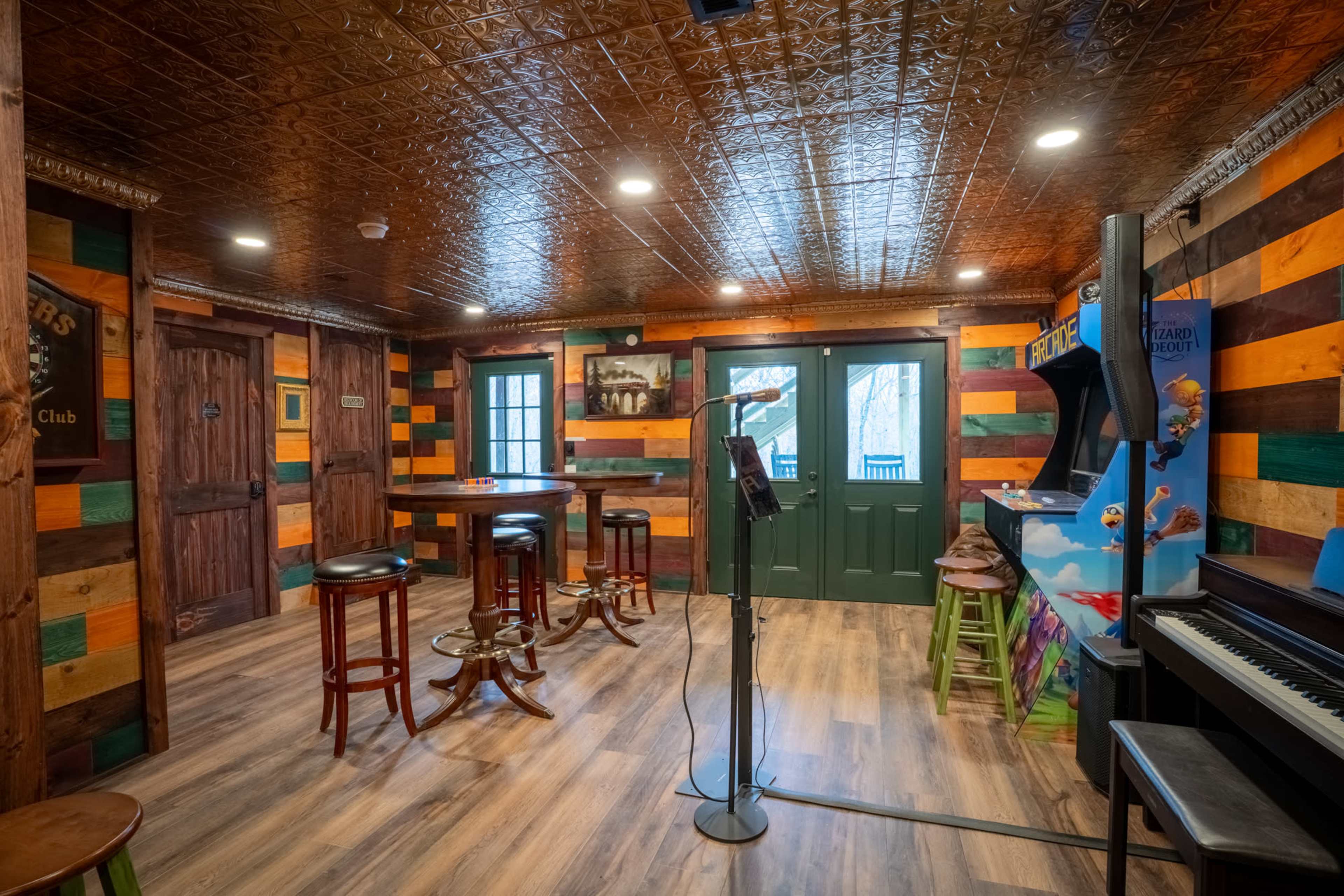 The image shows a rustic interior of a bar or club with wooden walls painted in various colors, tall tables, stools, and a karaoke machine.