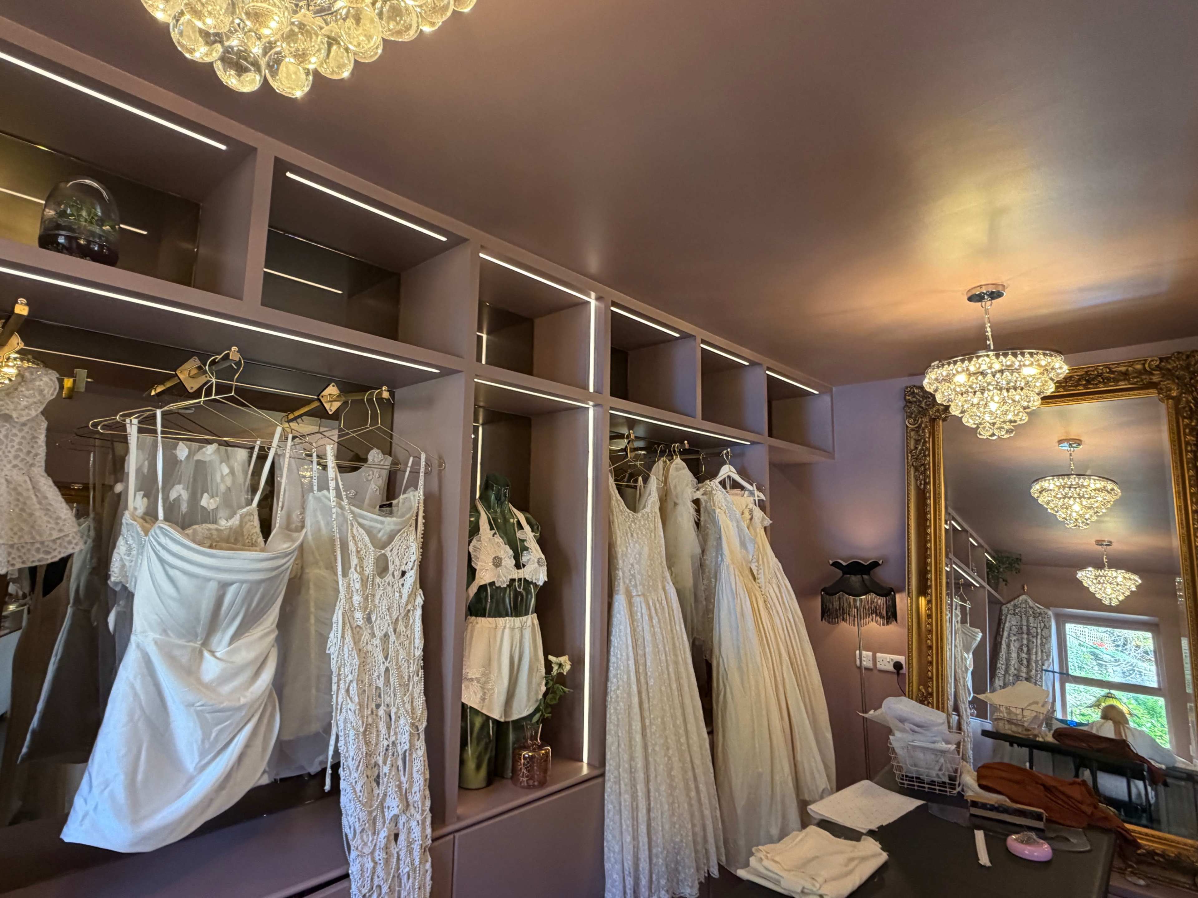 A boutique fitting room displays an array of white dresses hanging on racks, with a full-length mirror and chandeliers illuminating the space.